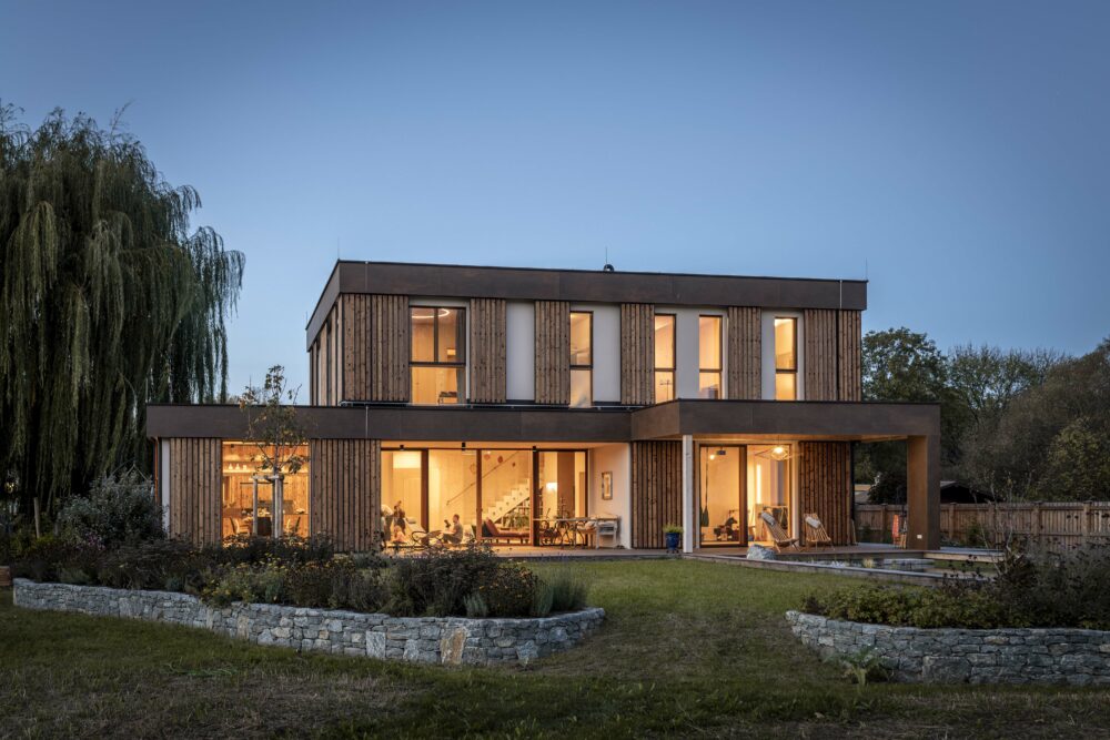 Modernes zweigeschossiges Wohnhaus mit vertikalen Holzlamellen aus Piz Bernina Lava, beleuchtetem Innenraum, großen Glasflächen und Natursteinmauern im Garten bei einsetzender Dämmerung.