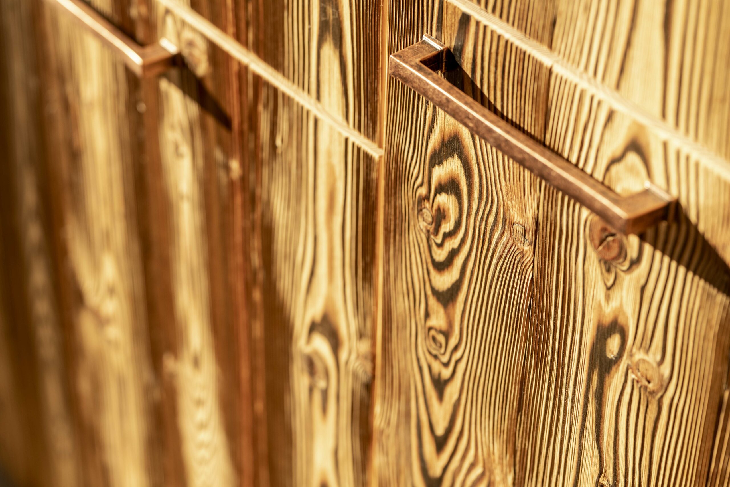 Nahaufnahme einer Küchenfront mit markanter Maserung und bronzefarbenem Griff, gefertigt aus Piz Bernina Lava – thermisch veredeltes Holz mit charakterstarker Struktur.