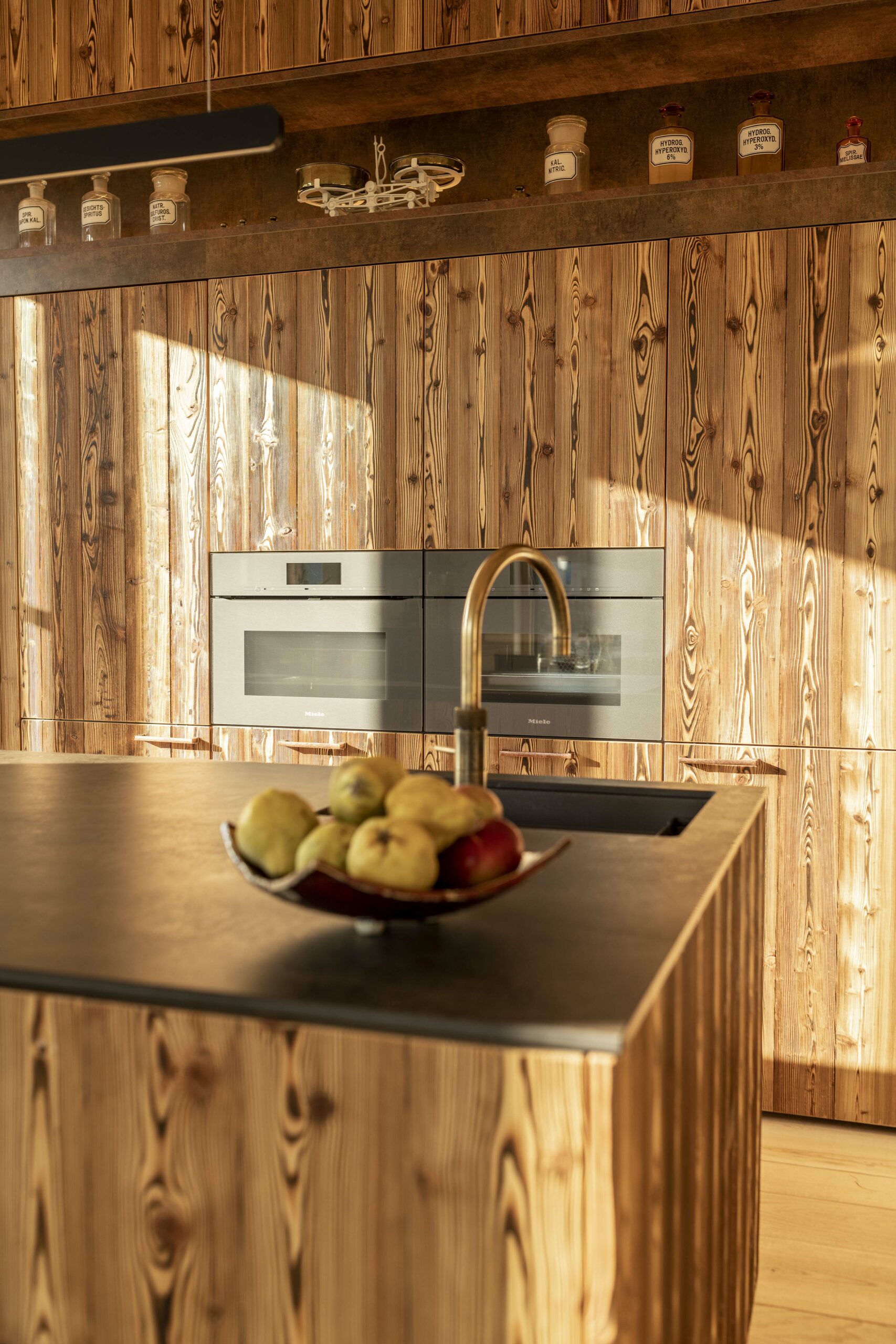 Moderne Küche mit dunkler Arbeitsplatte und Fronten aus thermisch behandeltem Holz Piz Bernina Lava – charaktervolle Maserung trifft auf stilvolle Geräteintegration.