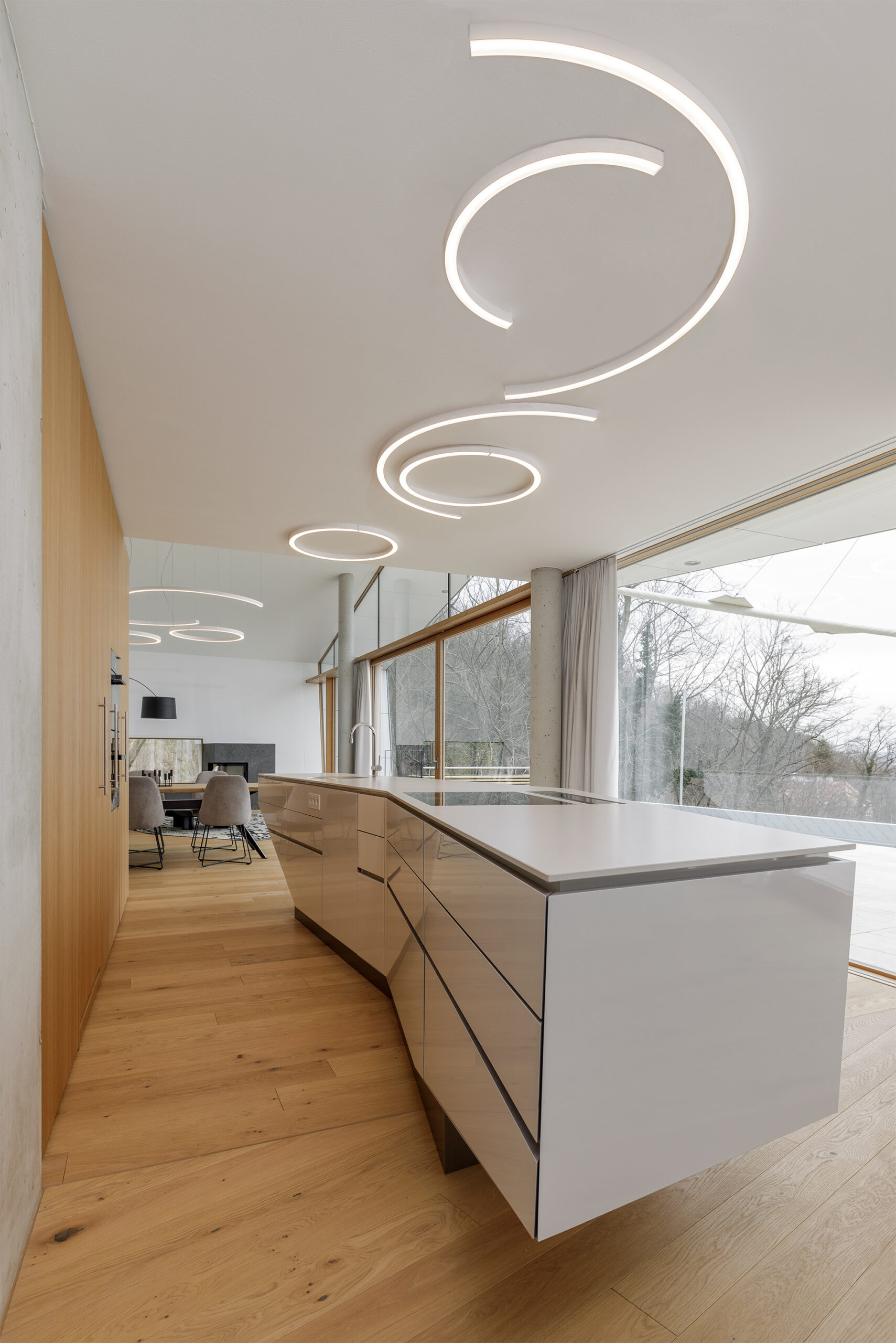 Moderne Küche mit weißer Designinsel, organischen Deckenleuchten und großzügiger Fensterfront mit Blick ins Freie, auf hellem Eichenboden Mareiner Simplon.