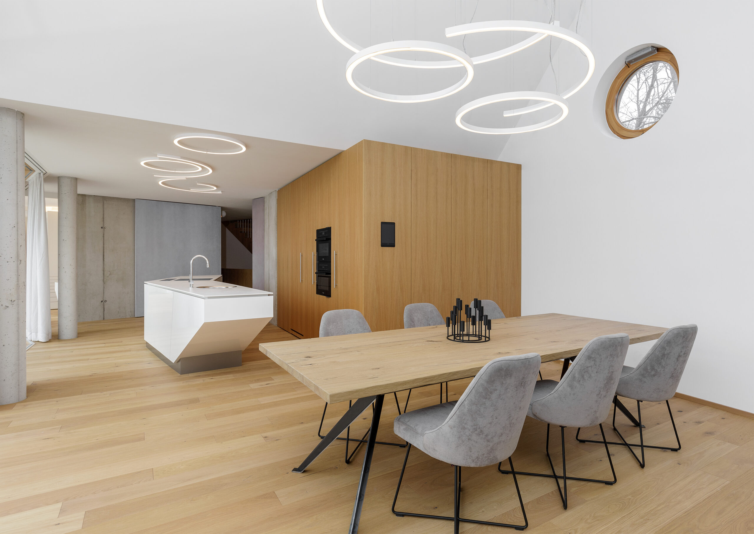 Wohnraum mit hellem Eichenboden (Mareiner Simplon) und Massivholztisch aus der Serie Crans Montana. Modernes Interieur mit klaren Linien, grauen Polsterstühlen und geometrischer Beleuchtung.