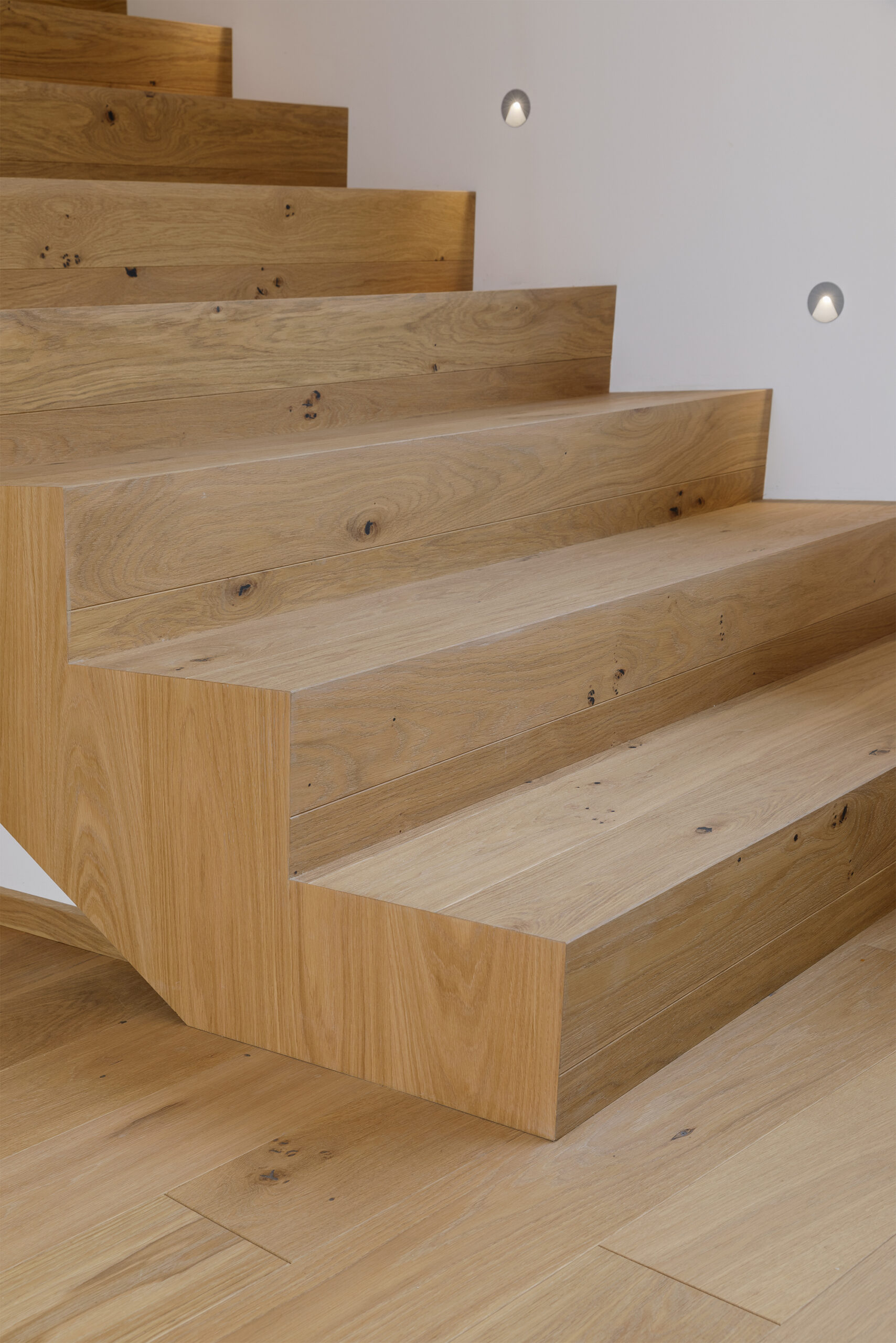 Nahaufnahme einer massiven Eichentreppe mit charaktervoller Maserung und Astlöchern aus Mareiner Simplon, flächenbündig verarbeitet und mit integrierter Wandbeleuchtung.