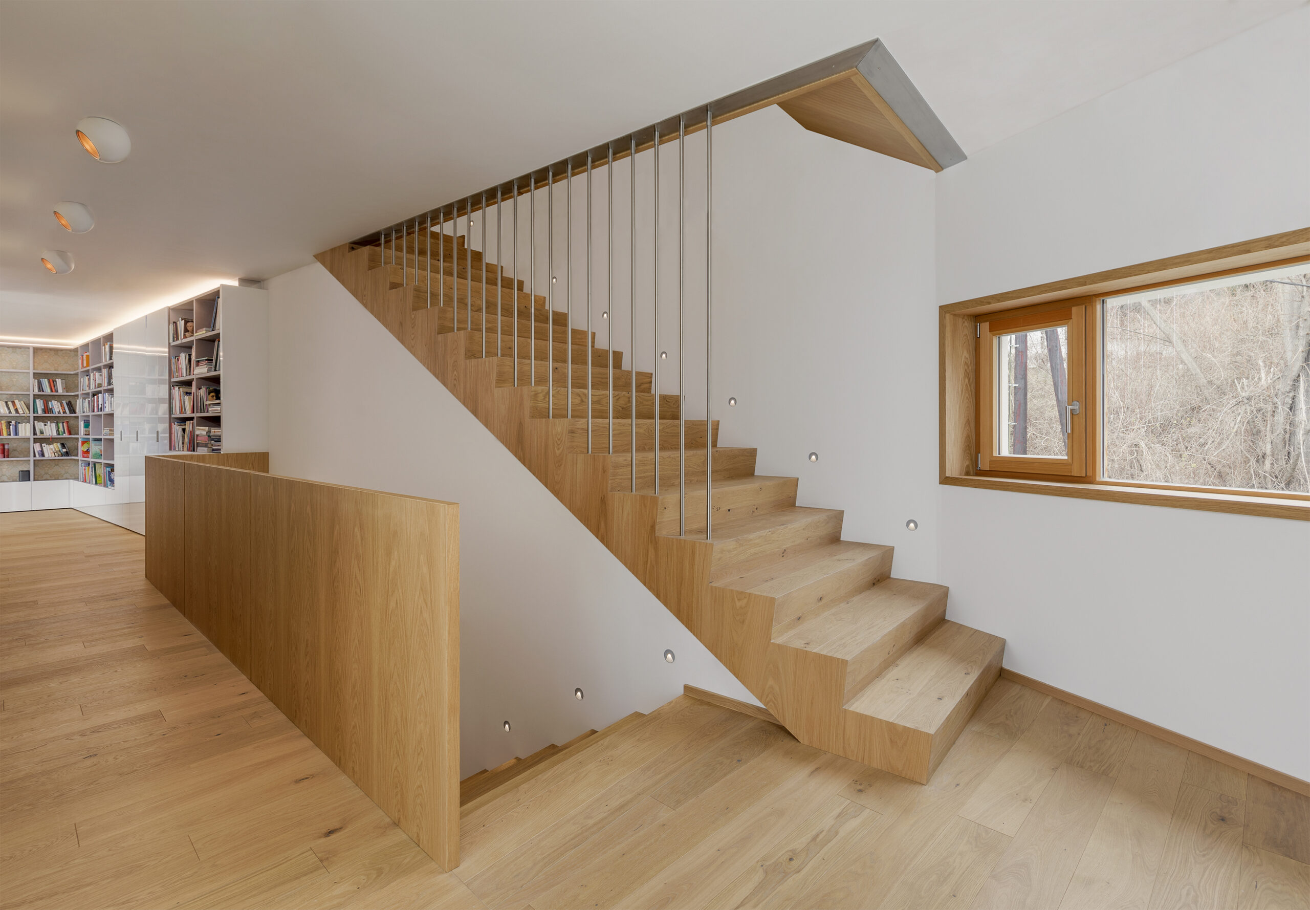 Moderner Treppenaufgang aus Mareiner Simplon Eiche mit schlankem Metallgeländer, daneben eine helle Bibliothek mit weißen Regalen, alles auf hellem Holzfußboden.