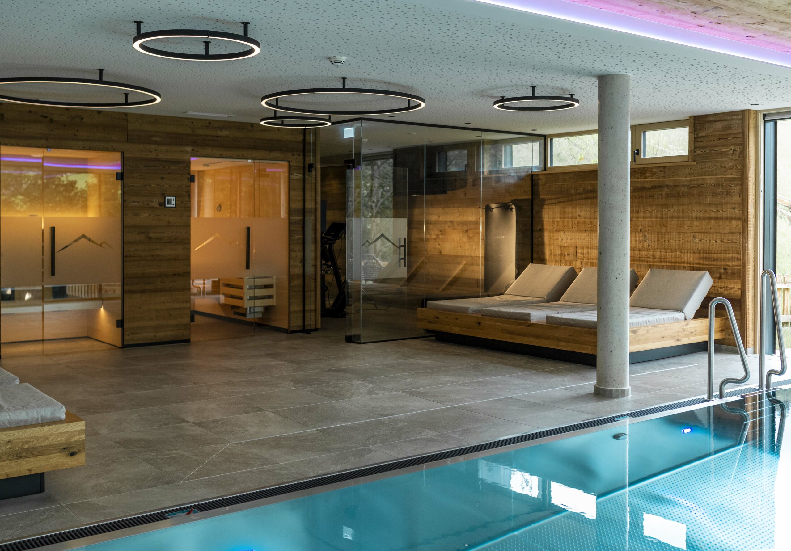 Eleganter Wellnessbereich mit Innenpool und Liegen, verkleidet mit Mareiner Piz Nair Holz im Bandsägeschnitt, kombiniert mit Glas- und Betonakzenten.