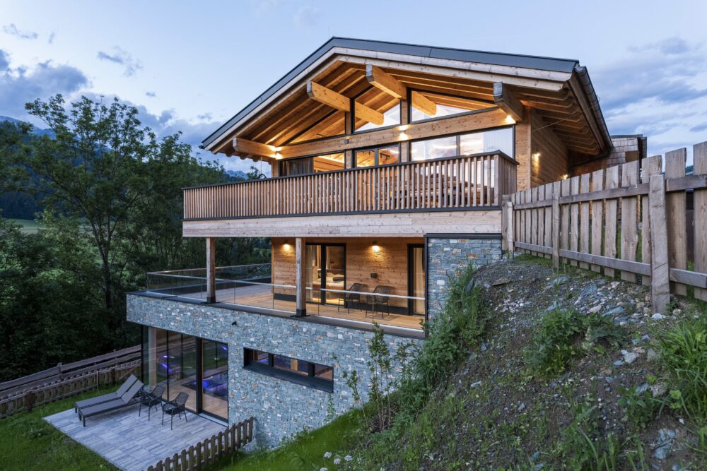 Modernes Chalet mit Fassade aus gehacktem Piz Bernina Holz und Naturstein-Sockel, großzügigen Glasflächen, Holzbalkonen und sichtbaren Dachbalken in alpiner Hanglage.