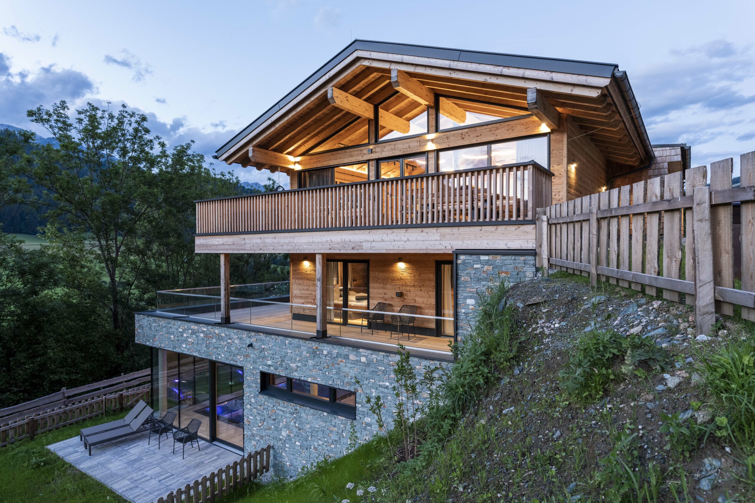 Modernes Chalet mit Fassade aus gehacktem Piz Bernina Holz und Naturstein-Sockel, großzügigen Glasflächen, Holzbalkonen und sichtbaren Dachbalken in alpiner Hanglage.
