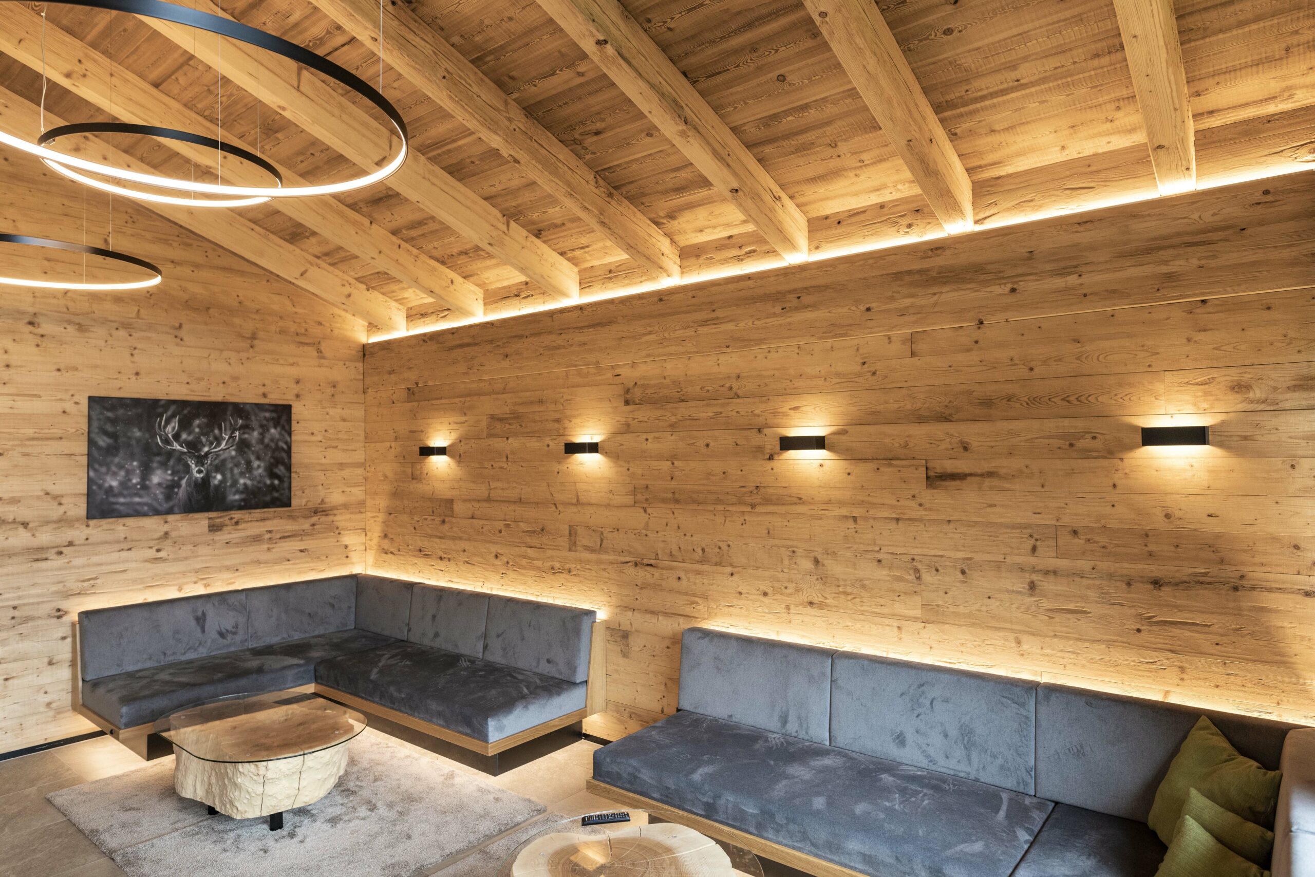 Gemütlicher Lounge-Bereich mit Wand- und Deckenverkleidung aus gehacktem Piz Bernina Holz, indirekter Beleuchtung, modernen Leuchten und Sitzbänken mit grauen Polstern.