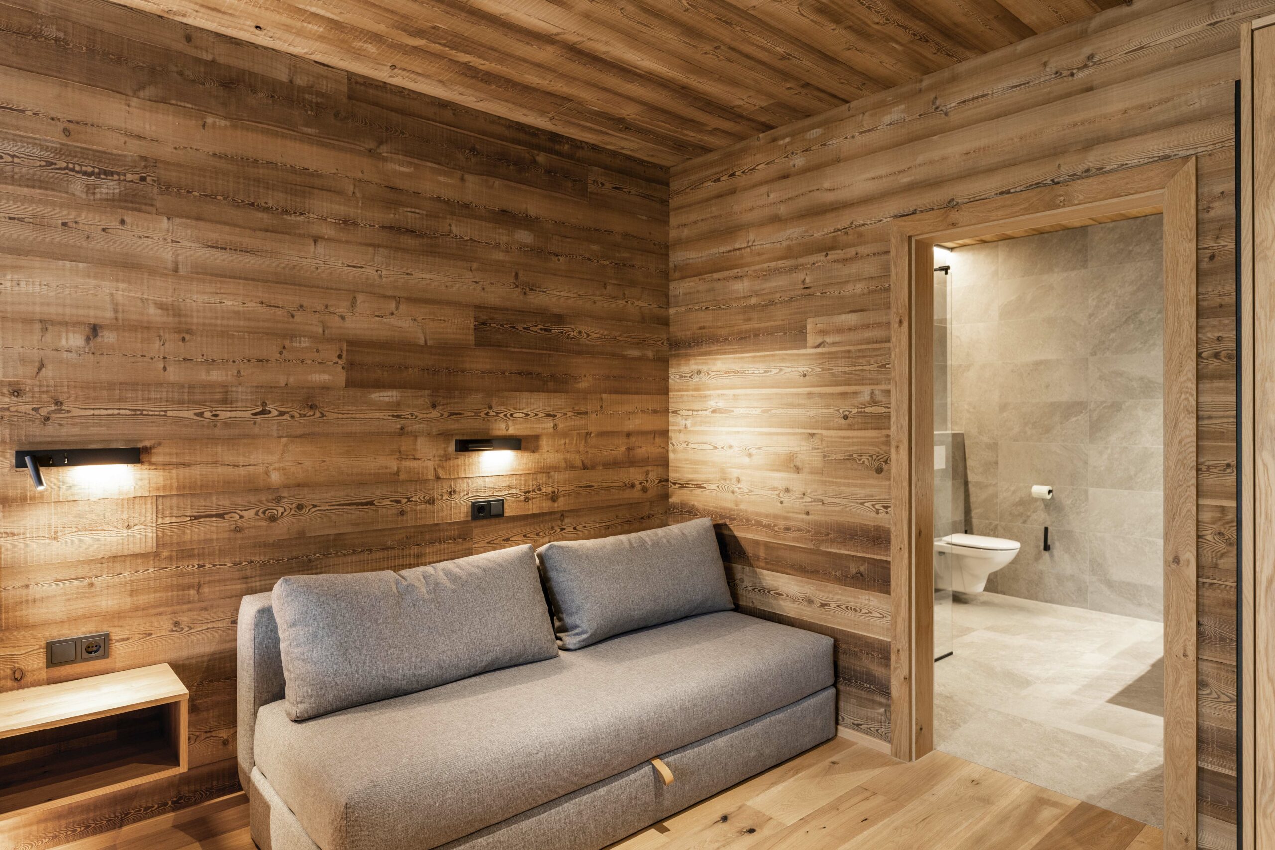 Modernes Zimmer mit Wand- und Deckenverkleidung aus Mareiner Marmolada Vecchia, kombiniert mit grauem Sofa und angrenzendem Bad in Natursteinoptik.