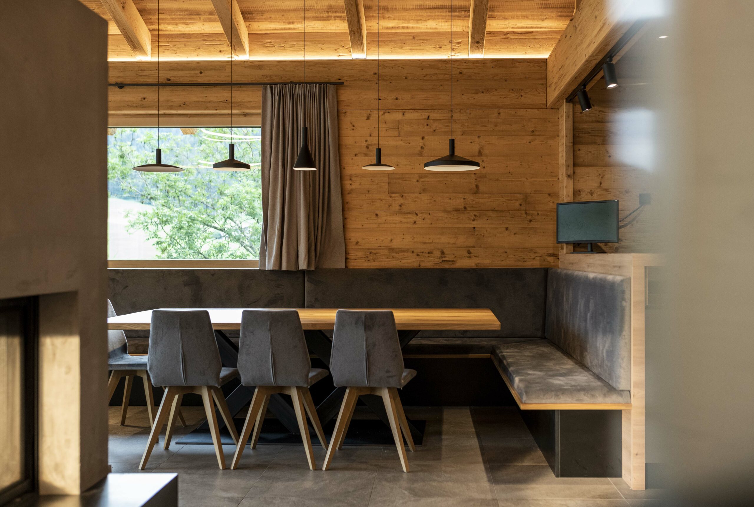 Moderner Essbereich mit Wandverkleidung in Piz Nair mit Bandsägeschnitt – charaktervoll strukturiertes Holz in warmem Farbton, kombiniert mit grauer Polsterbank, Natursteinboden und eleganter Beleuchtung.