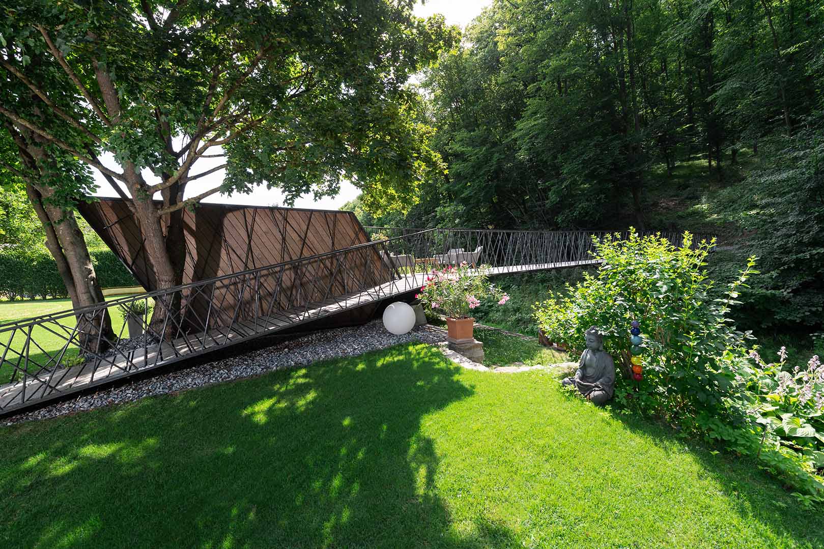 Gesamtansicht des Fußgängerstegs in Rosenburg mit Kebony-Terrassendielen und filigranem Metallgeländer. Die Konstruktion integriert sich harmonisch in die grüne Gartenlandschaft mit Buddha-Statue und Blüten.