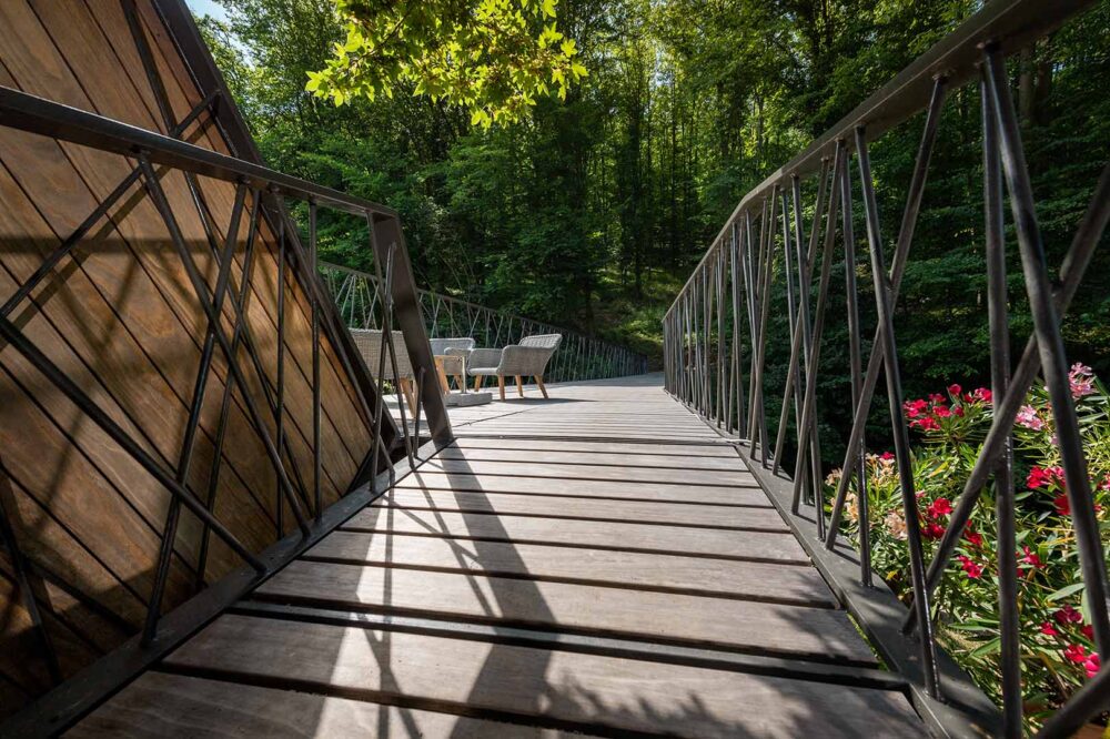 Holzsteg mit Kebony-Terrassendielen und schwarzen Metallgeländern führt durch einen bewaldeten Bereich. Links moderne Holzverkleidung, rechts blühende Sträucher. Am Ende des Stegs laden Sessel zum Verweilen ein.