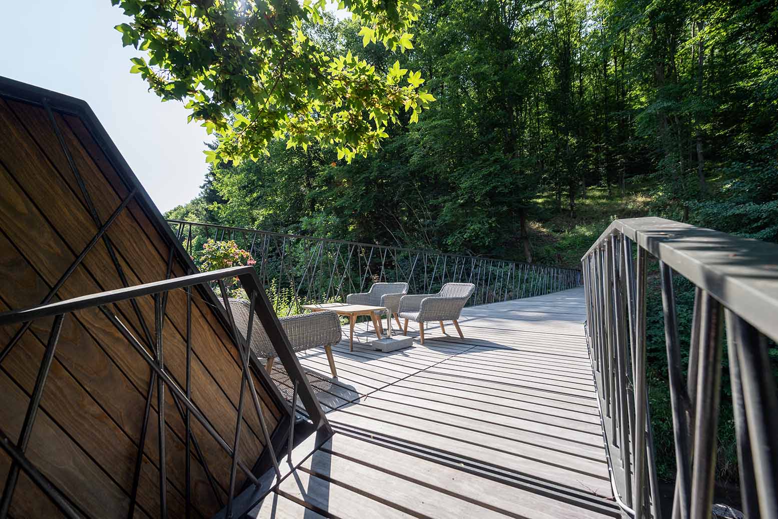 Brücke mit Kebony-Terrassendielen in Rosenburg, seitlich eingefasst mit filigranen Metallgeländern. Im Vordergrund diagonale Holzverkleidung, im Hintergrund gemütlicher Sitzbereich mit Sesseln und Tisch, umgeben von grünem Wald.