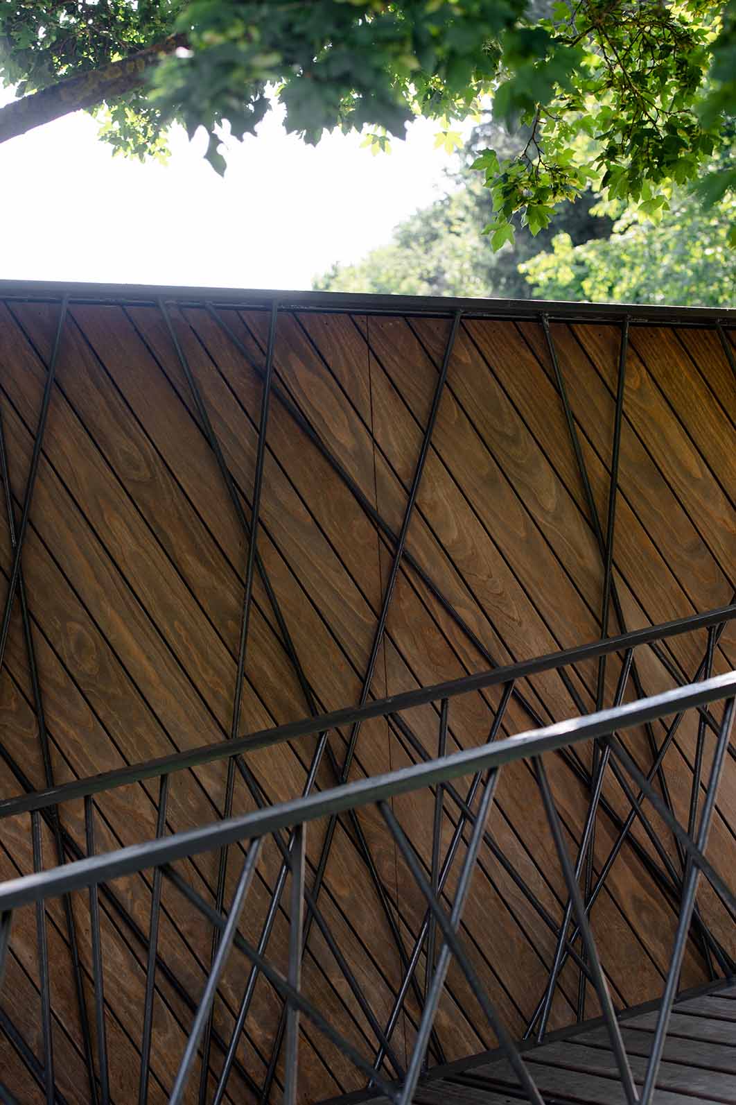 Nahaufnahme der Wandverkleidung des Fußgängerstegs in Rosenburg mit schräg verlegten Kebony-Dielen und filigraner schwarzer Stahlstruktur im geometrischen Muster, darunter ein Handlauf und im oberen Bereich belaubte Äste.