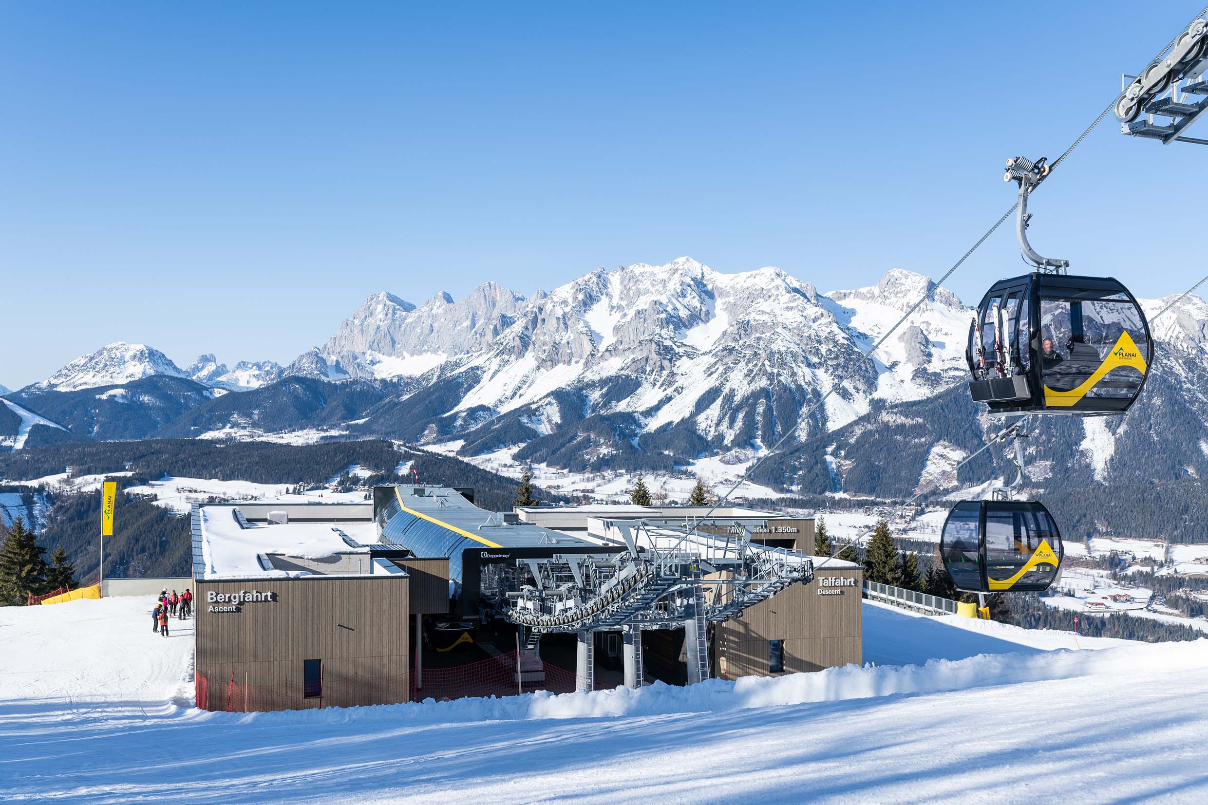 Weitwinkelaufnahme einer modernen Seilbahn-Bergstation mit vertikaler Holzfassade aus Mareiner Piz Nair. Die gelb-schwarzen Gondeln schweben vor einer eindrucksvollen Winterkulisse mit schneebedeckten Gipfeln. Im Vordergrund präparierte Skipisten und wartende Wintersportler.