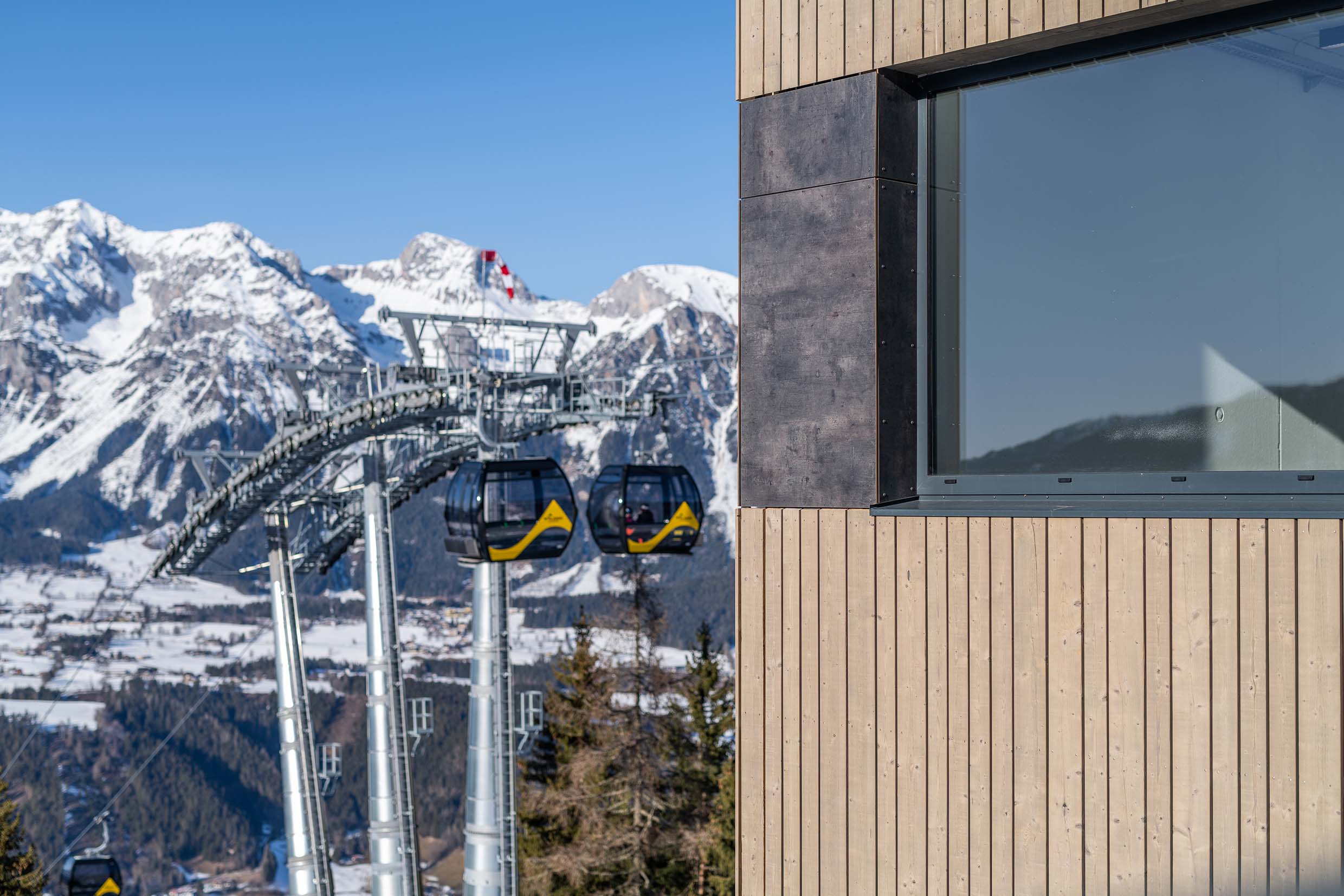 Moderne vertikale Holzfassade aus Mareiner Piz Nair mit dunkel gerahmtem Fenster. Im Hintergrund: eine Seilbahnstation mit Gondeln sowie verschneite Alpen. Die Holzstruktur ist klar erkennbar und steht im Kontrast zur schwarzen Fensterumrahmung und den schroffen Bergen.