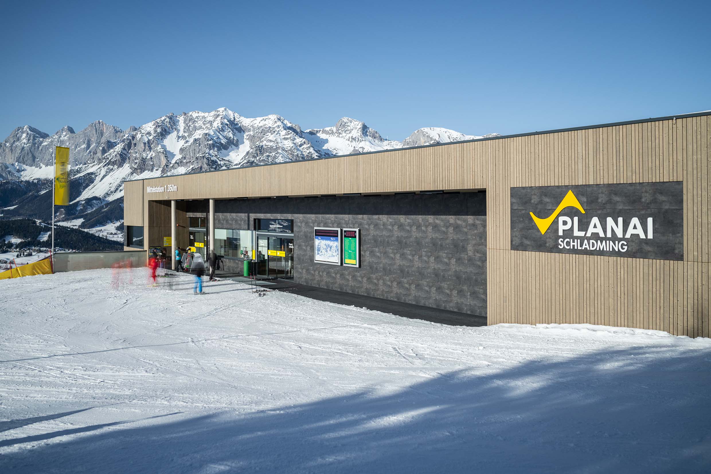 Architekturansicht der Planai-Mittelstation auf 1.350 m mit vertikaler Holzfassade aus Mareiner Piz Nair und dunklen Fassadenplatten. Im Vordergrund bewegen sich Skifahrer über die beschneite Fläche, im Hintergrund das imposante Dachsteinmassiv.