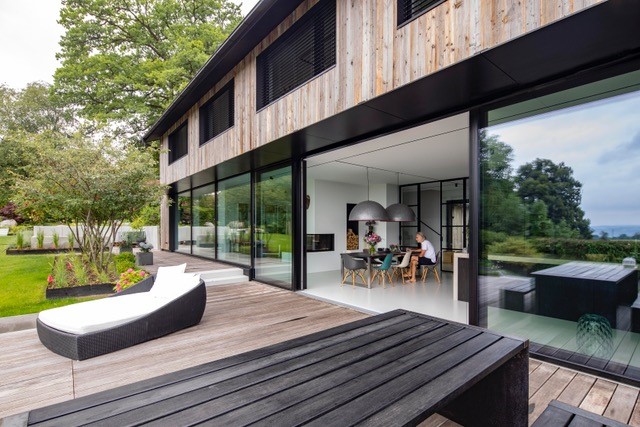 Modernes Wohnhaus mit vertikaler Zermatt-Holzfassade von Mareiner und großflächigen Glas-Schiebetüren zur Terrasse. Die Holzterrasse schafft fließende Übergänge zwischen Innen- und Außenraum, ergänzt durch moderne Outdoor-Möbel.