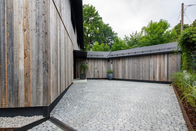 Einfahrt zu einem modernen Wohnhaus mit vertikal montierter Zermatt-Holzfassade von Mareiner. Der Vorplatz ist mit Natursteinpflaster belegt, umrahmt von schlichten Pflanzgefäßen und grüner Bepflanzung.