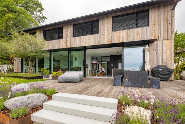 Rückansicht eines modernen Wohnhauses mit vertikaler Zermatt-Holzfassade von Mareiner. Die große Terrasse besteht aus Holzdielen, eingerahmt von blühenden Beeten und Natursteinen. Großflächige Fensterfronten öffnen den Wohnbereich zum Garten.