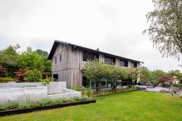 Zweigeschossiges Wohnhaus mit vertikaler Holzfassade aus Mareiner Zermatt. Das Gebäude steht in einem gepflegten Garten mit Rasenfläche, Hochbeeten und Bäumen. Die graubraune Holzstruktur der Fassade verleiht dem Haus eine natürliche, elegante Anmutung.
