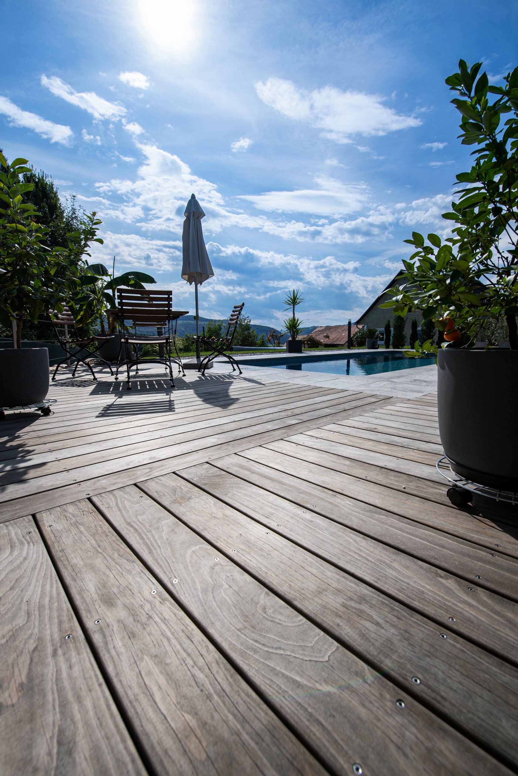 Großzügige Terrasse mit diagonal verlegtem Kebony-Holz in eleganter Vergrauung, Blick auf Pool, Gartenmöbel und üppige Pflanzen. Die tiefstehende Sonne erzeugt markante Schatten und verleiht der Szene sommerliche Atmosphäre.