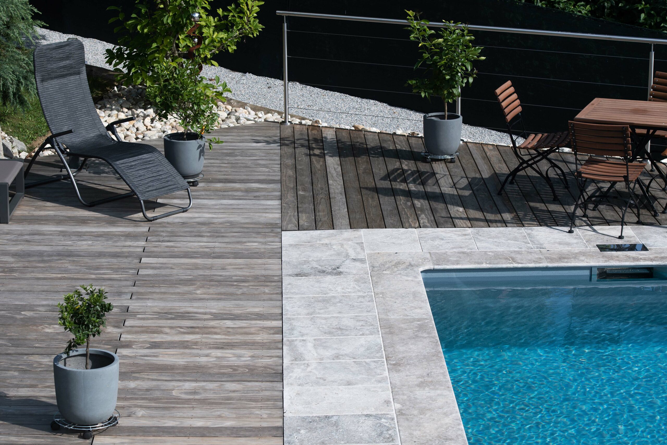 Moderne Poollandschaft mit graubraun patiniertem Kebony-Terrassenholz, angrenzendem Naturstein-Poolrand und Sitz- sowie Liegemöbeln. Pflanzkübel mit Zitrusbäumchen setzen Akzente, ein Metallgeländer begrenzt die Fläche.