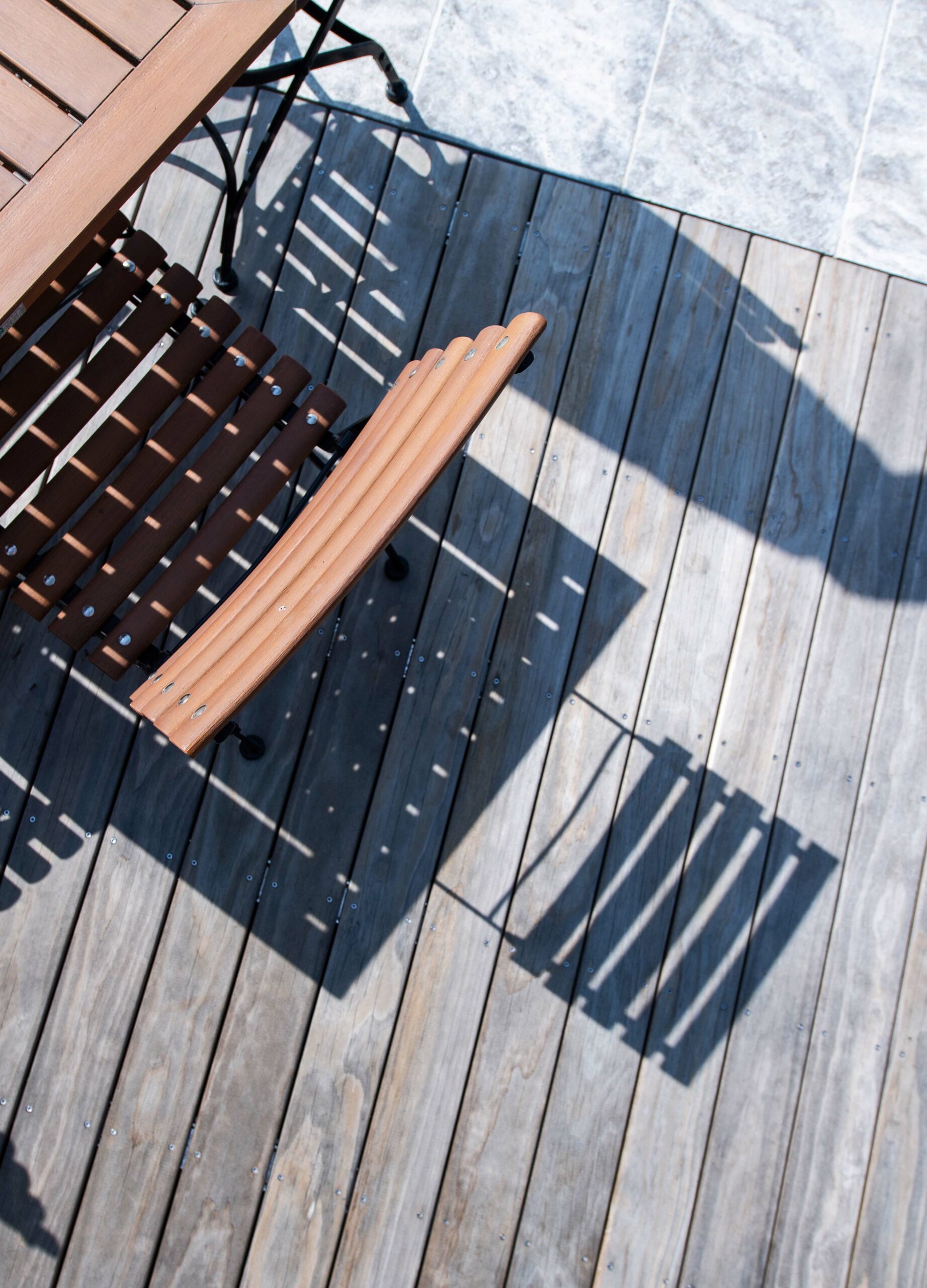 Nahaufnahme einer Kebony-Holzterrasse mit sichtbarer silbergrauer Patina. Eine hölzerne Gartenstuhllehne und Tischkante werfen markante Schatten auf die Dielen. Im oberen Bildbereich sind hellgraue Natursteinplatten am Poolrand zu sehen.