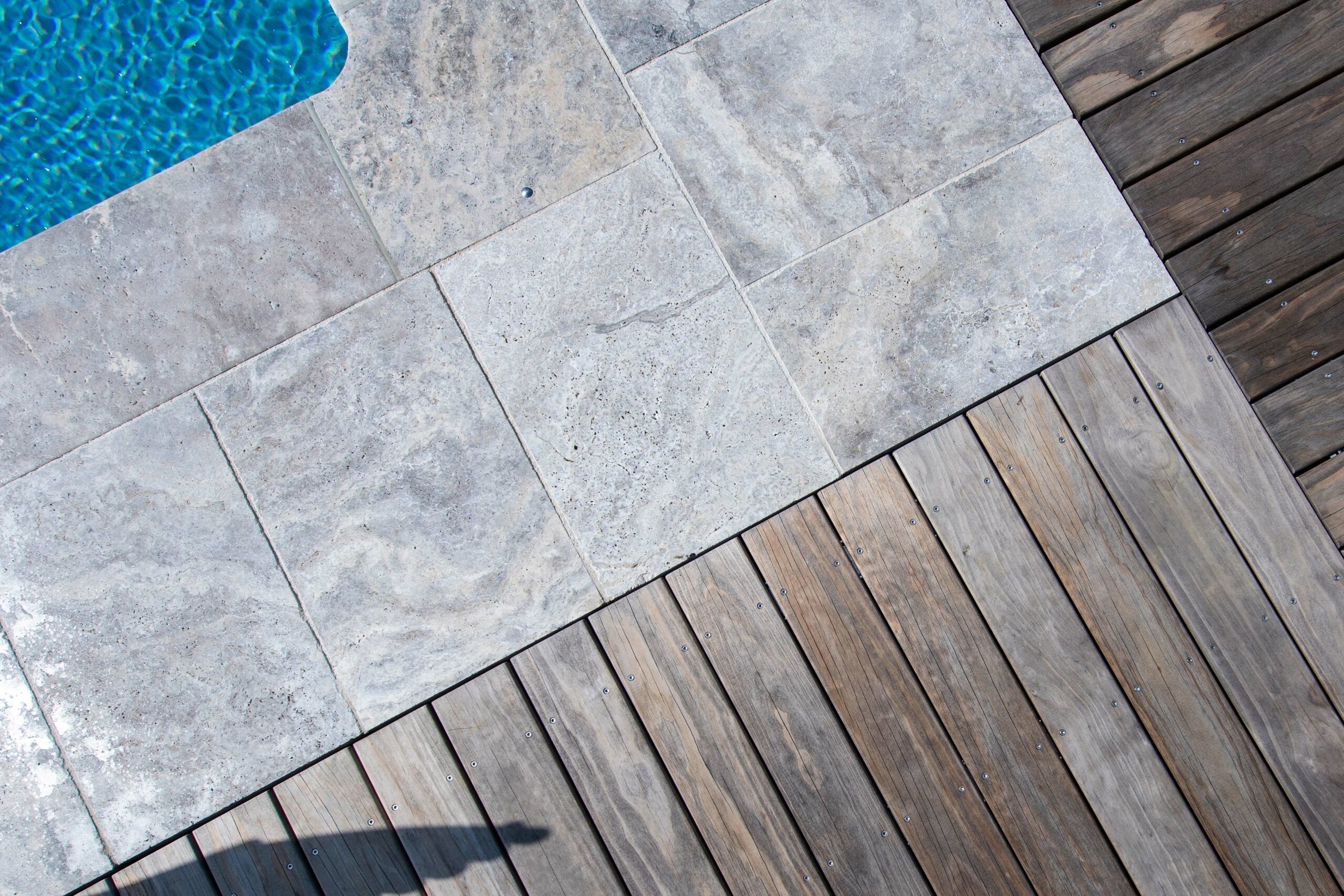 Nahaufnahme einer Terrasse mit diagonal verlegtem Kebony-Holzdeck und angrenzender grauer Natursteinumrandung eines Pools. Das klare Wasser reflektiert das Sonnenlicht, die Materialien zeigen eine elegante, wettergegerbte Patina.