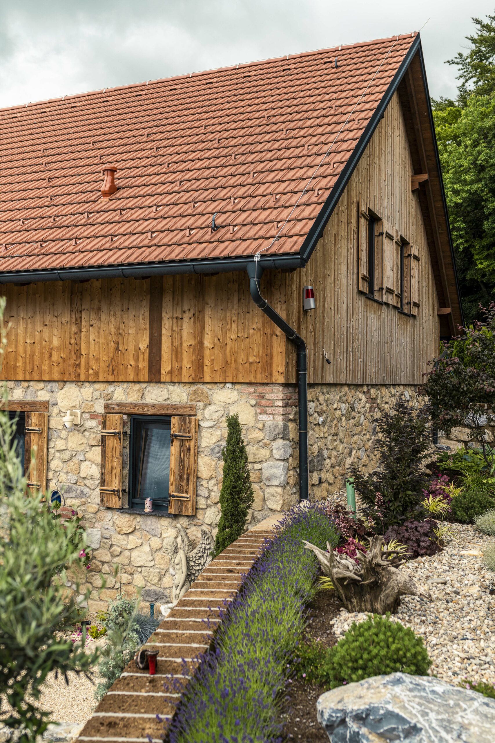 Traditionelle Hausfassade mit vertikaler Holzverkleidung aus Gran Paradiso im Obergeschoss und rustikaler Natursteinmauer im Sockelbereich. Vor dem Haus ein liebevoll gestalteter Garten mit Lavendel, Ziergehölzen und einer kleinen Engelsfigur.