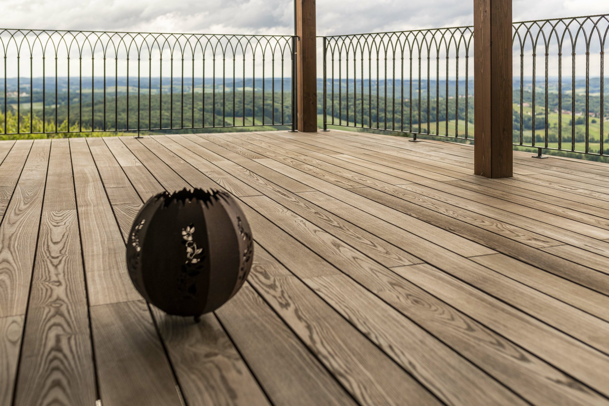 Weitläufige Terrasse mit edlem Boden aus Gardasee-Holz. Im Vordergrund steht eine dekorative schwarze Metallkugel. Die filigrane Metallbalustrade gibt den Blick auf eine hügelige Landschaft und weiten Horizont frei.