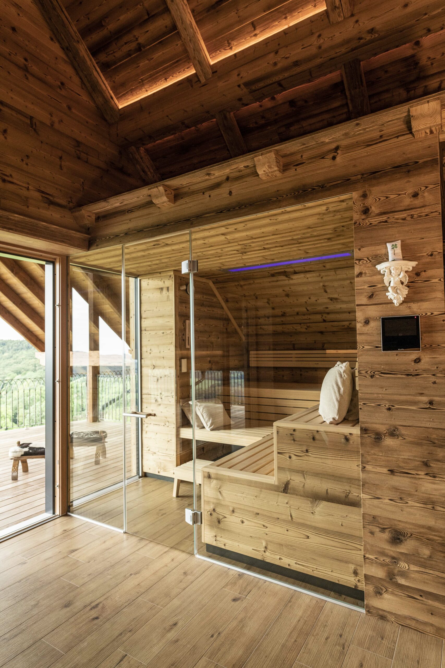 Modern interpretierter alpiner Wellnessbereich mit Sauna aus Marmolada Lava Holz. Die Glasfront gibt den Blick auf eine Terrasse mit Talpanorama frei. Sichtbar sind rustikale Balken, warme Beleuchtung und eine gemütlich gestaltete Saunabank mit Kissen.