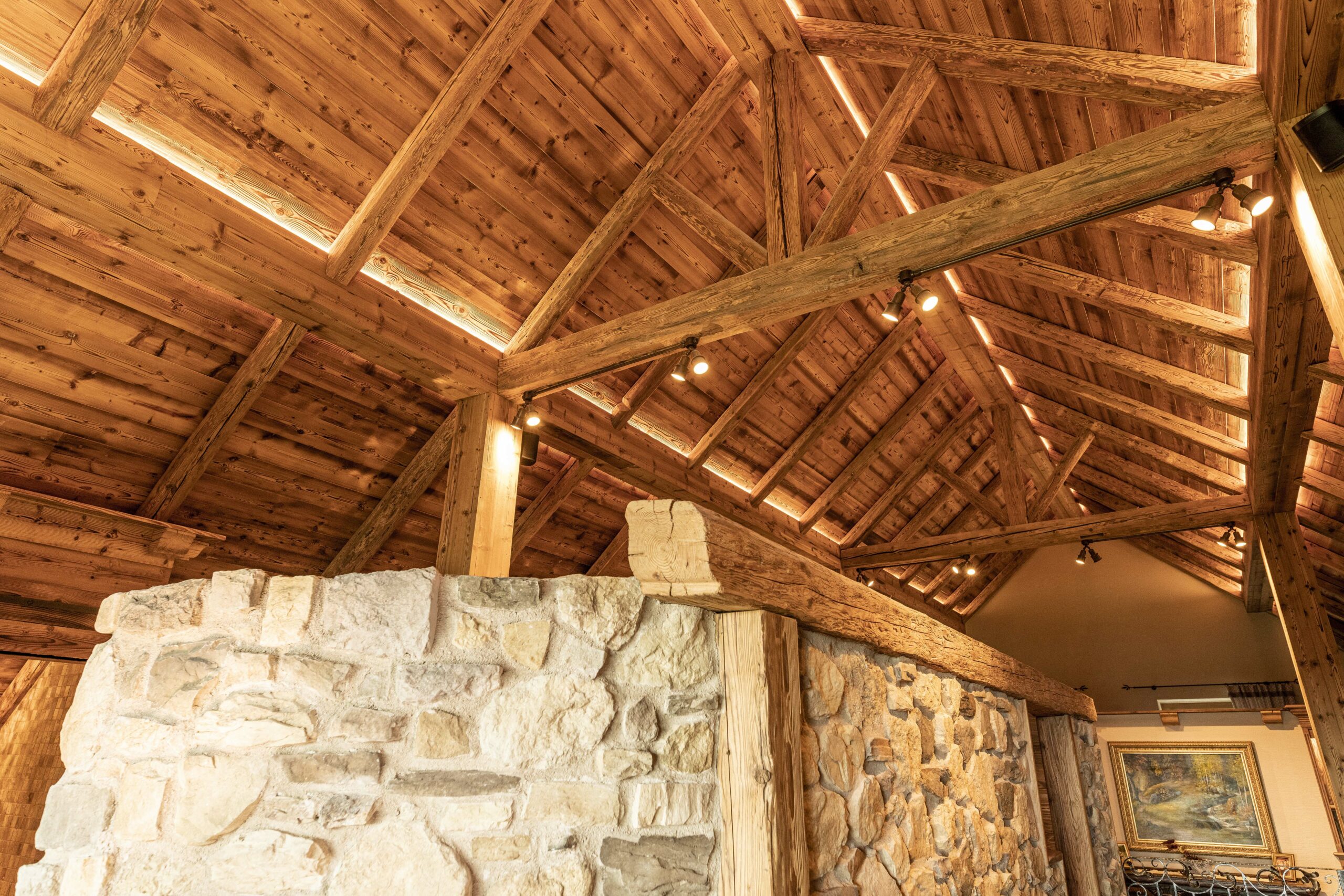 Detailaufnahme eines rustikalen Sichtdachstuhls aus Marmolada Lava Holz mit sichtbaren Trägern und stimmungsvoller Beleuchtung. Darunter eine massive Mauer aus Naturstein, im Hintergrund ein gerahmtes Landschaftsbild an der Wand.