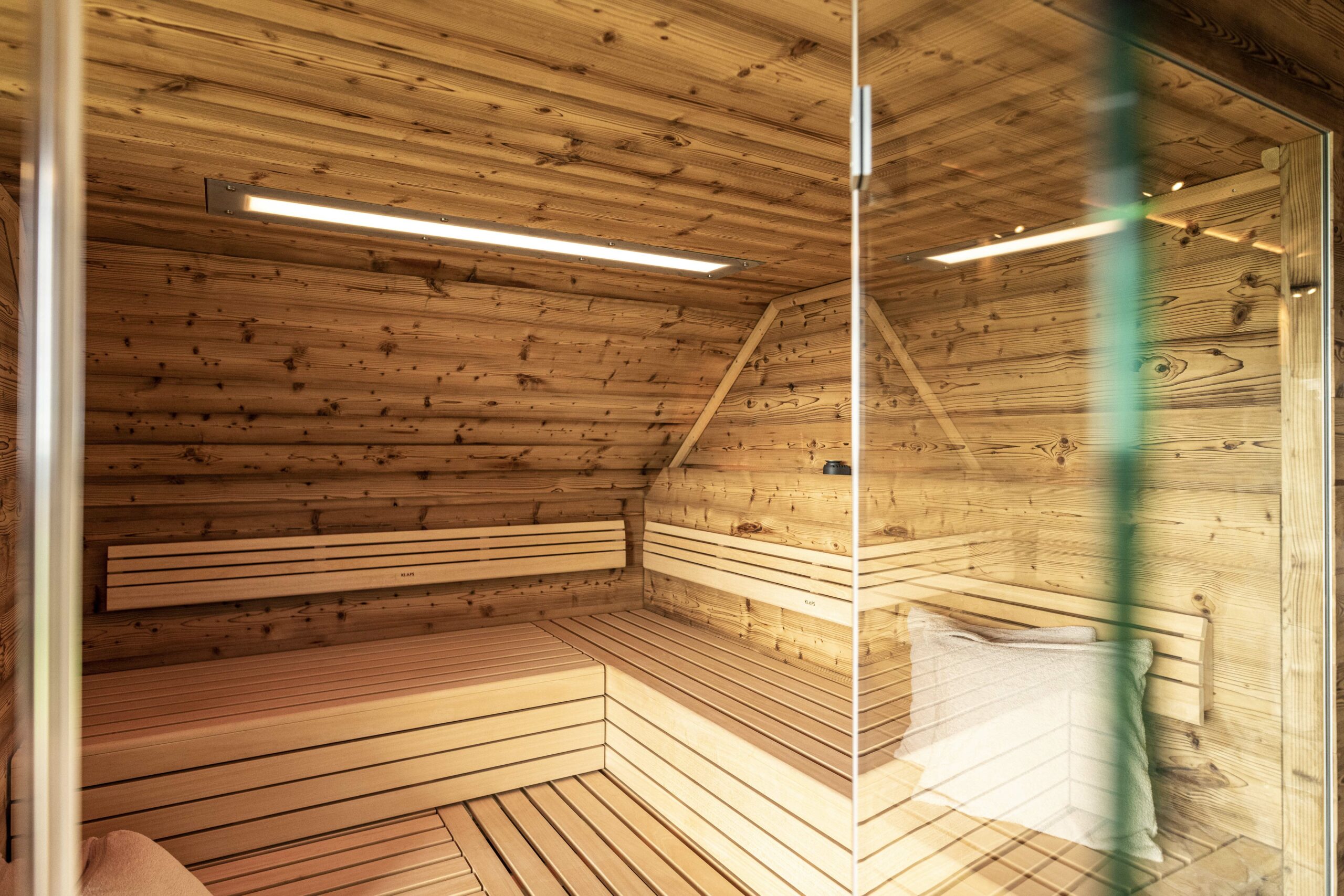 Hochwertige Sauna mit Wand- und Deckenverkleidung aus Marmolada Lava Holz. Die Innenbänke aus hellem Holz kontrastieren harmonisch mit der rustikalen, astreichen Struktur der Verkleidung. Sichtbar ist eine Glaswand sowie ein Saunakissen.