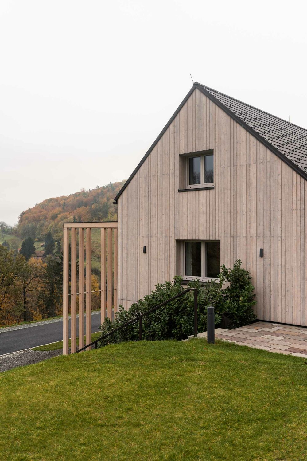 Giebelansicht eines modernen Hauses mit Fassade aus vorvergrautem Mareiner Piz Nair in vertikaler Schalung. Im Vordergrund eine gepflegte Grünfläche und eine Pergola aus schlanken Holzlamellen, im Hintergrund bewaldete Hügellandschaft in Herbstfarben.