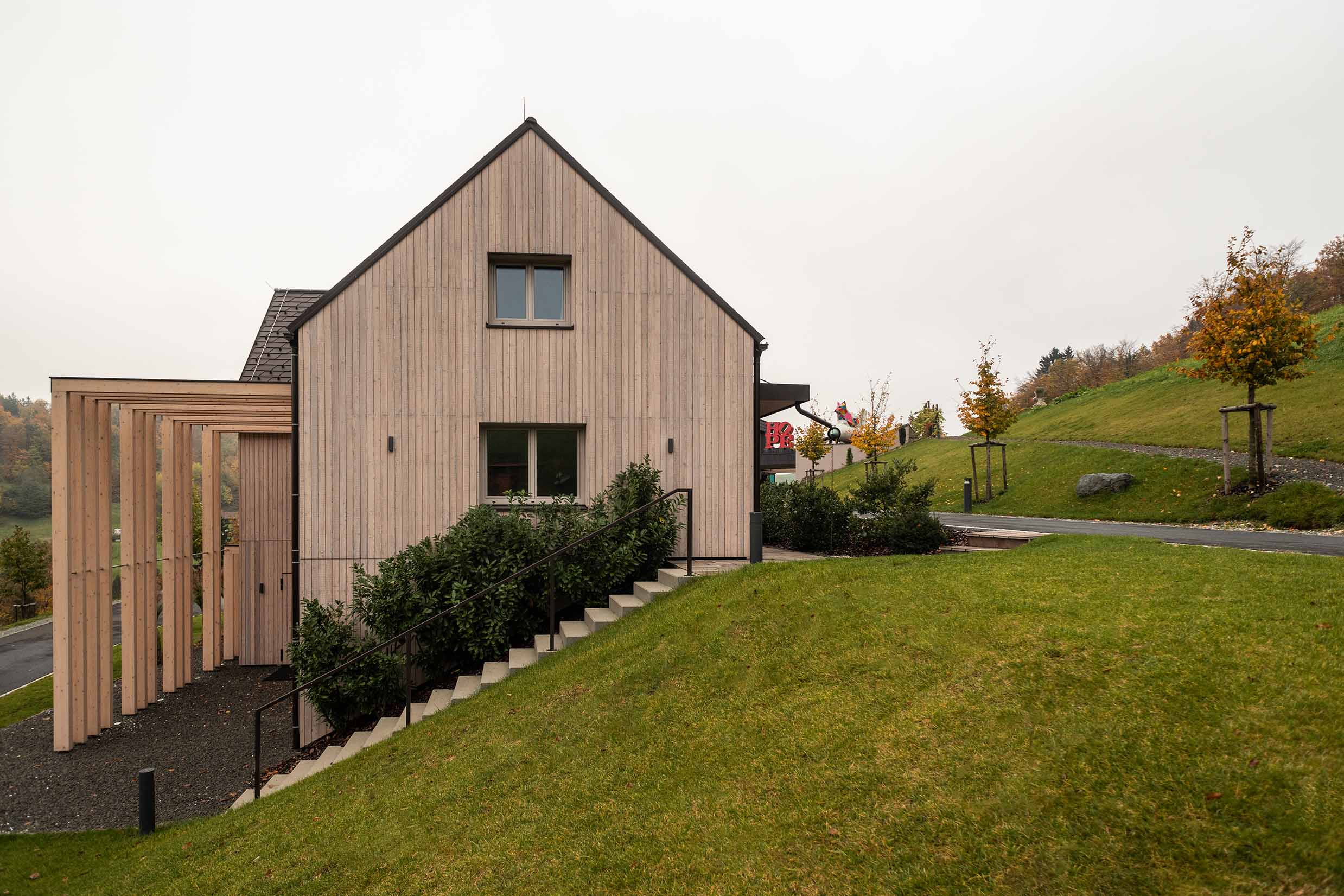 Seitenansicht eines modernen Hauses in Hanglage mit vertikaler Holzfassade aus vorvergrautem Mareiner Piz Nair. Sichtbar sind eine Pergola aus hohen Holzlamellen, ein seitlicher Aufgang mit Geländer sowie ein gepflegter Garten mit herbstlicher Bepflanzung.