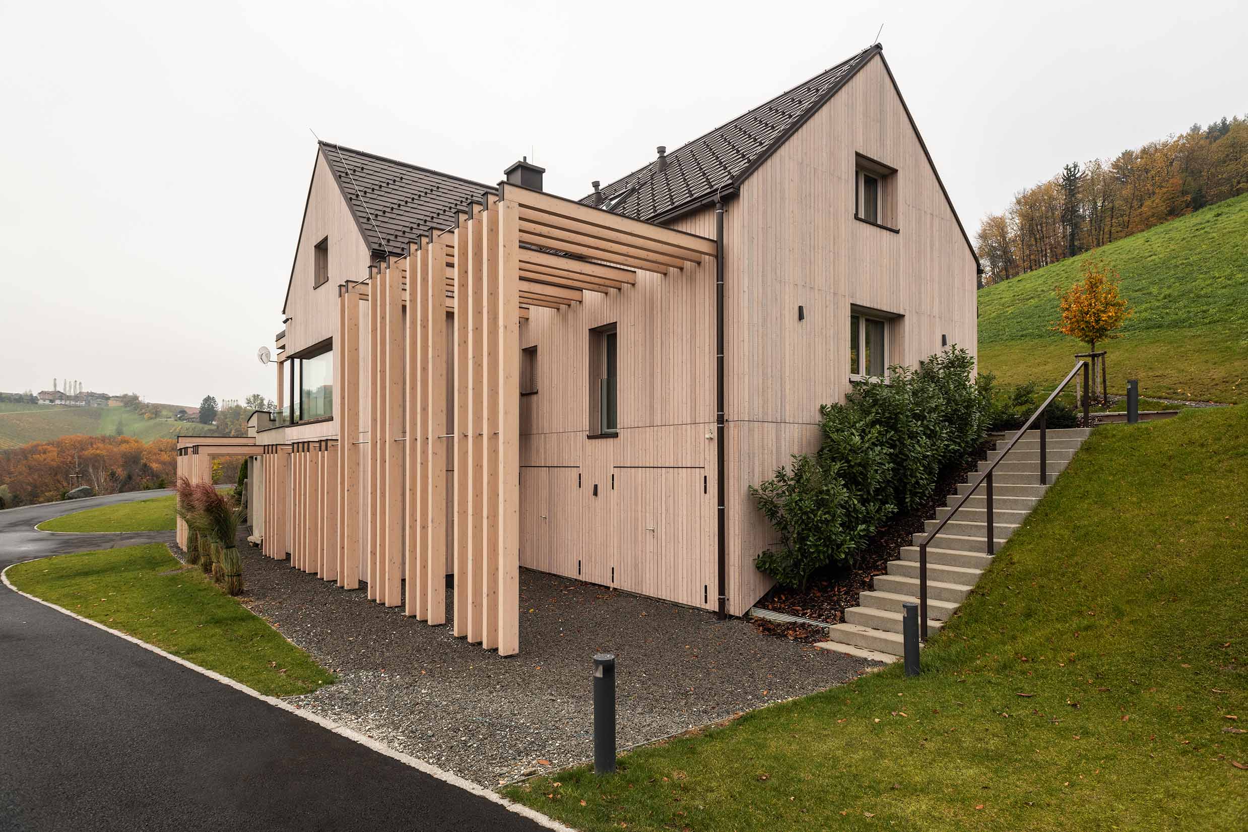 Architektonisch modern gestaltetes Haus in Hanglage, mit vertikaler Holzfassade aus vorvergrautem Mareiner Piz Nair. Vor dem Gebäude schmale, hohe Holzelemente als Sichtschutz oder Pergola. Umgeben von Wiesen, Hügellandschaft und herbstlichem Laub.
