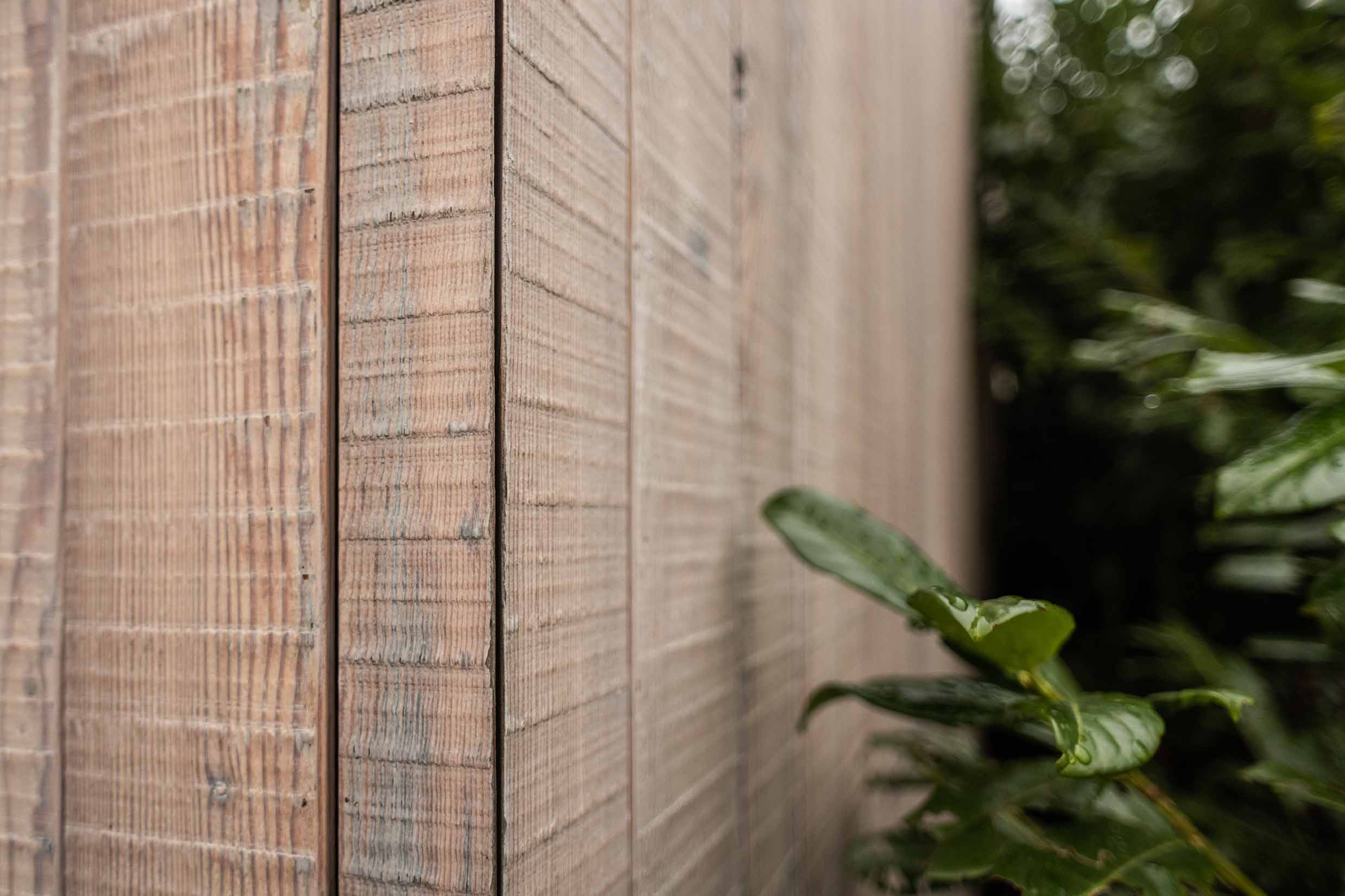 Nahaufnahme einer Holzfassade aus Mareiner Piz Nair, vorvergraut und im charakteristischen Bandsägeschnitt ausgeführt. Die raue, strukturierte Oberfläche des Holzes ist klar erkennbar; im Vordergrund unscharf ein nasses Blatt eines Zierstrauchs.