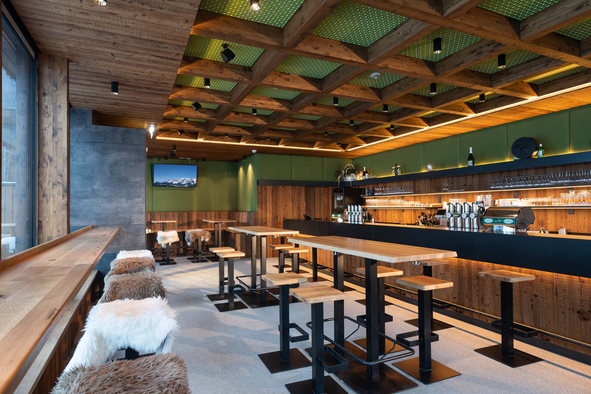 Modern interpretierter Barraum mit markanter Wand- und Deckenverkleidung aus Gran Paradiso 212°. Die dunkle Holzstruktur kombiniert sich mit grünen Akustikelementen in der Kassettendecke, einer eleganten Theke, Hocker mit Fellauflagen und stimmungsvoller Beleuchtung.