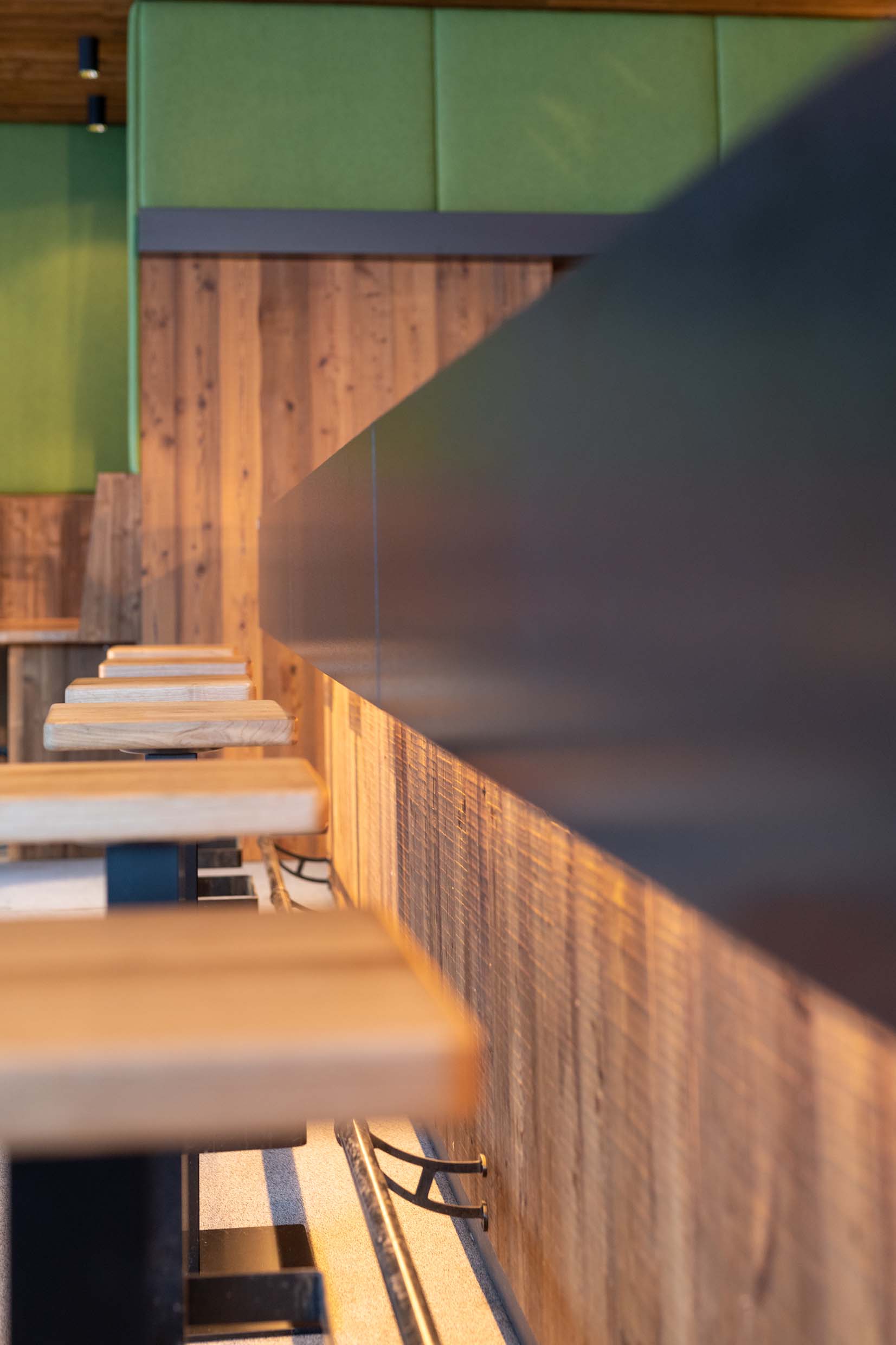 Stimmungsvolle Nahaufnahme entlang einer Bartheke, verkleidet mit Gran Paradiso 212°-Holz im Bandsägeschnitt. Im Vordergrund sind Hocker mit Holzsitzen zu sehen, dahinter grüne Akustikpaneele und die warme Lichtstimmung des Innenraums.