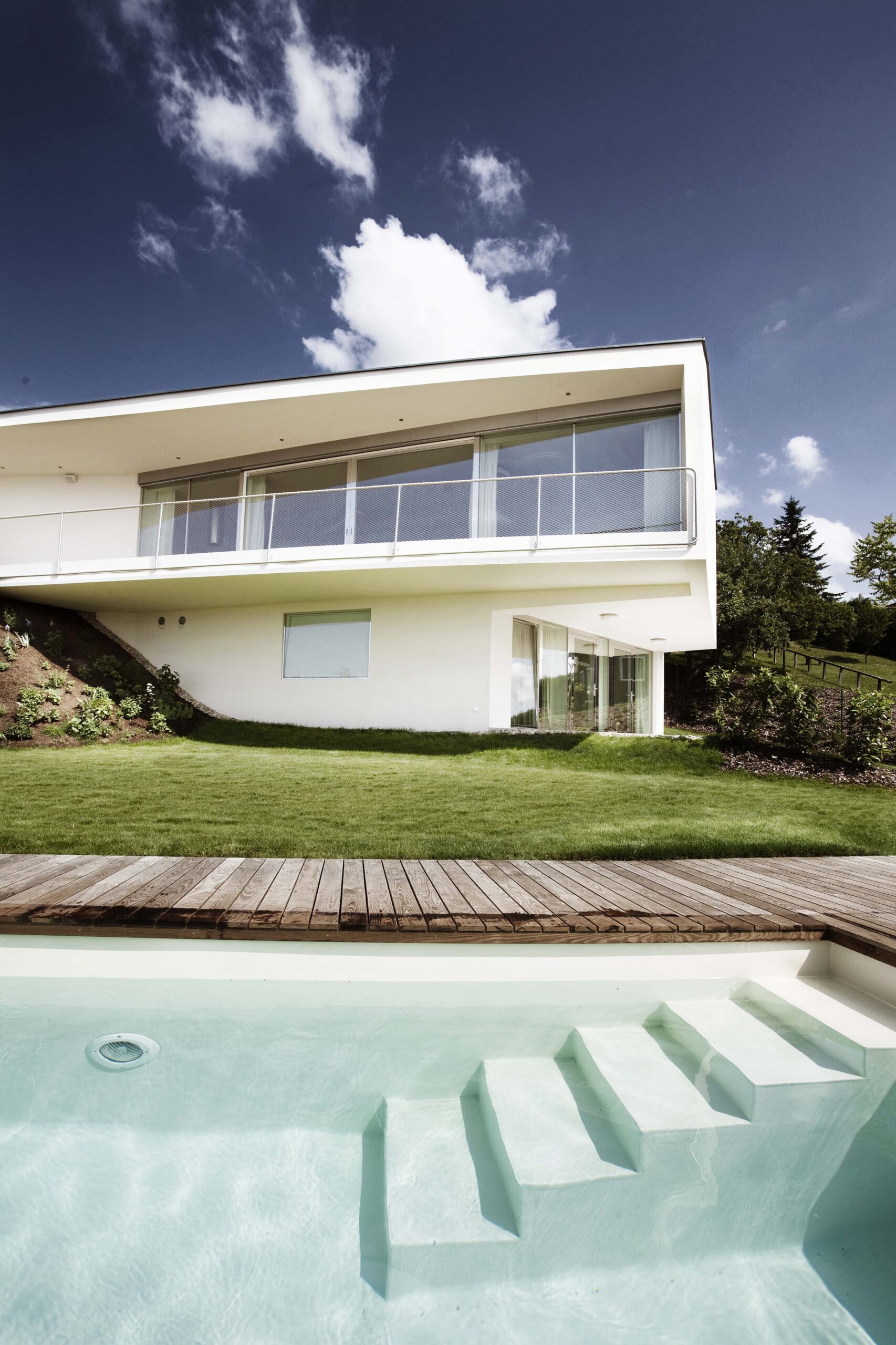 Blick vom Pool auf eine moderne Villa mit Holzterrasse aus Mareiner Gardasee. Die klare Architektur des Hauses ergänzt das natürliche Ambiente aus Wasser, Rasen und Holz unter blauem Himmel mit einzelnen Wolken.