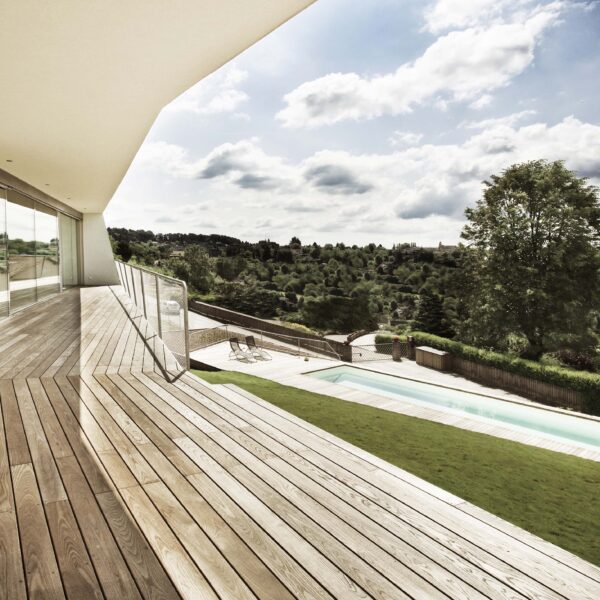 Spacious terrace made of Mareiner Gardasee wood with glass front and view of the pool and green hilly landscape. The clear lines of the architecture contrast with the natural surroundings under a partly cloudy sky.