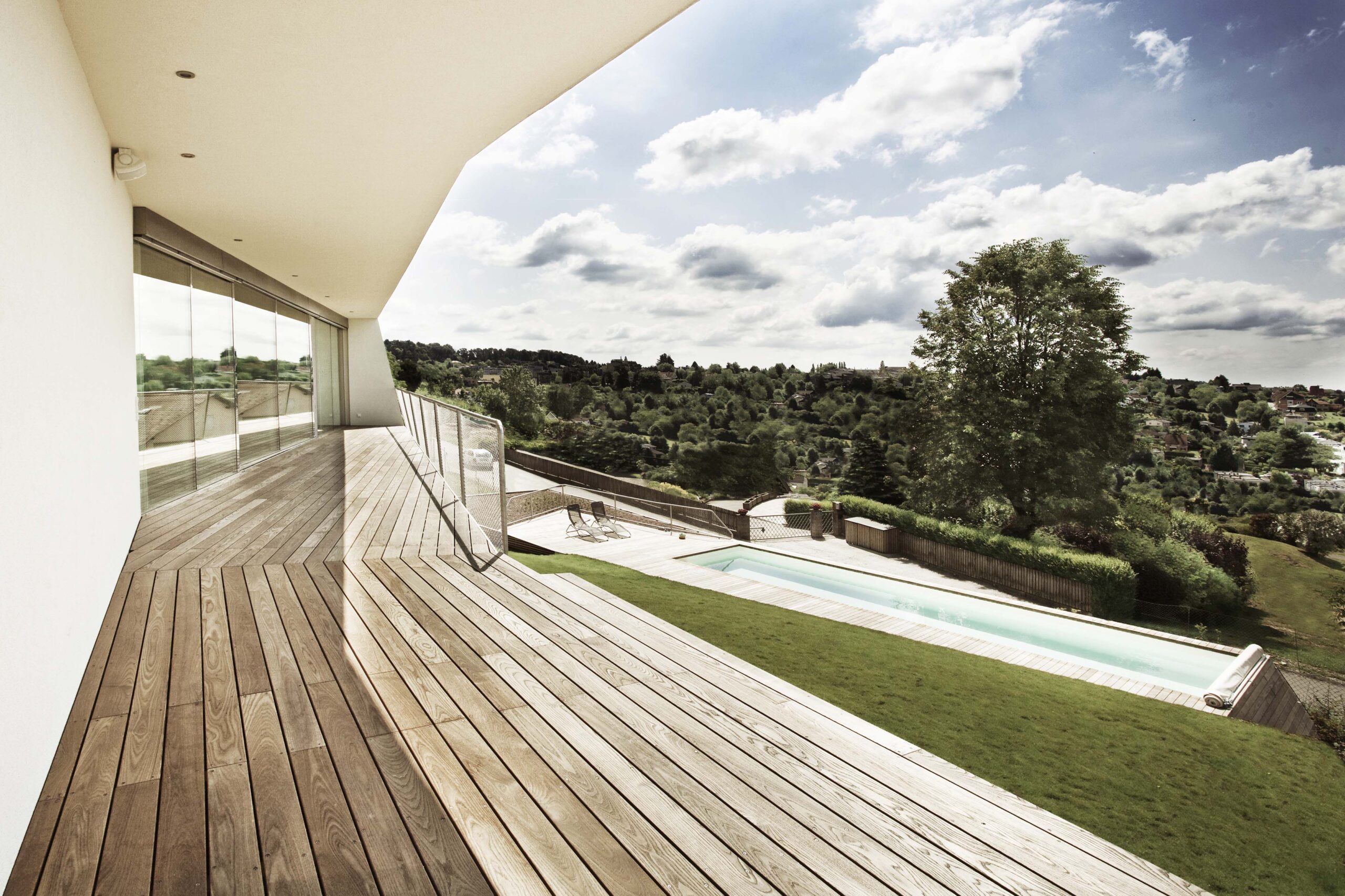 Weitläufige Terrasse aus Mareiner Gardasee Holz mit Glasfront und Ausblick auf Pool und grüne Hügellandschaft. Die klare Linienführung der Architektur kontrastiert mit der natürlichen Umgebung unter teils bewölktem Himmel.