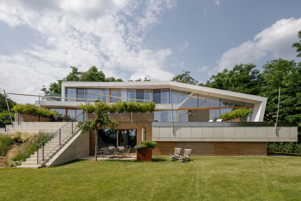 Moderne Villa mit großzügigen Glasflächen und einer markanten, geometrisch gestalteten Dachform. Die Fassade kombiniert helle Putz- und Plattenelemente mit vertikalen Holzlamellen. Die großzügige Terrasse ist mit Begrünung und Glasgeländern versehen. Darunter liegt ein ebenerdiger Wohnbereich mit direktem Zugang zum gepflegten Garten.