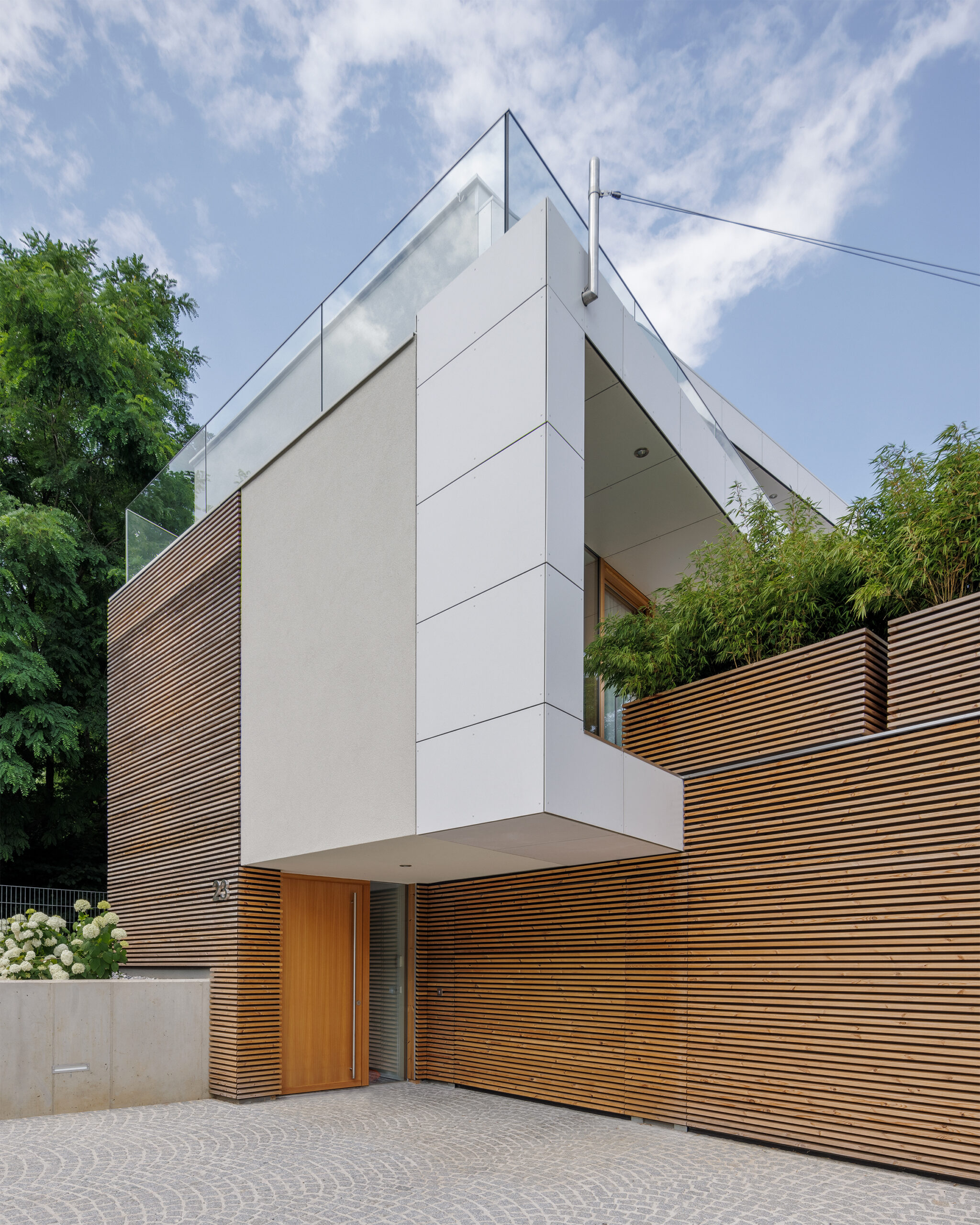 Moderne Hausfassade mit einer Kombination aus vertikalen Holzlamellen, glatten weißen Fassadenplatten und Glasgeländern auf der Dachterrasse. Der Eingangsbereich ist überdacht, mit einer Holztür und Pflanztrögen mit Bambus im oberen Bereich.