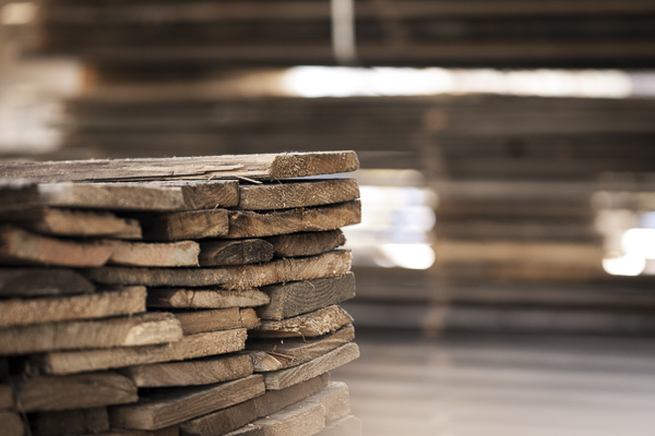 Gestapelte, unbehandelte Holzbohlen in einem Lager von Mareiner. Die grob zugeschnittenen Bretter sind unregelmäßig aufgeschichtet und warten auf die weitere Verarbeitung. Im Hintergrund sind weitere Holzstapel unscharf zu erkennen.