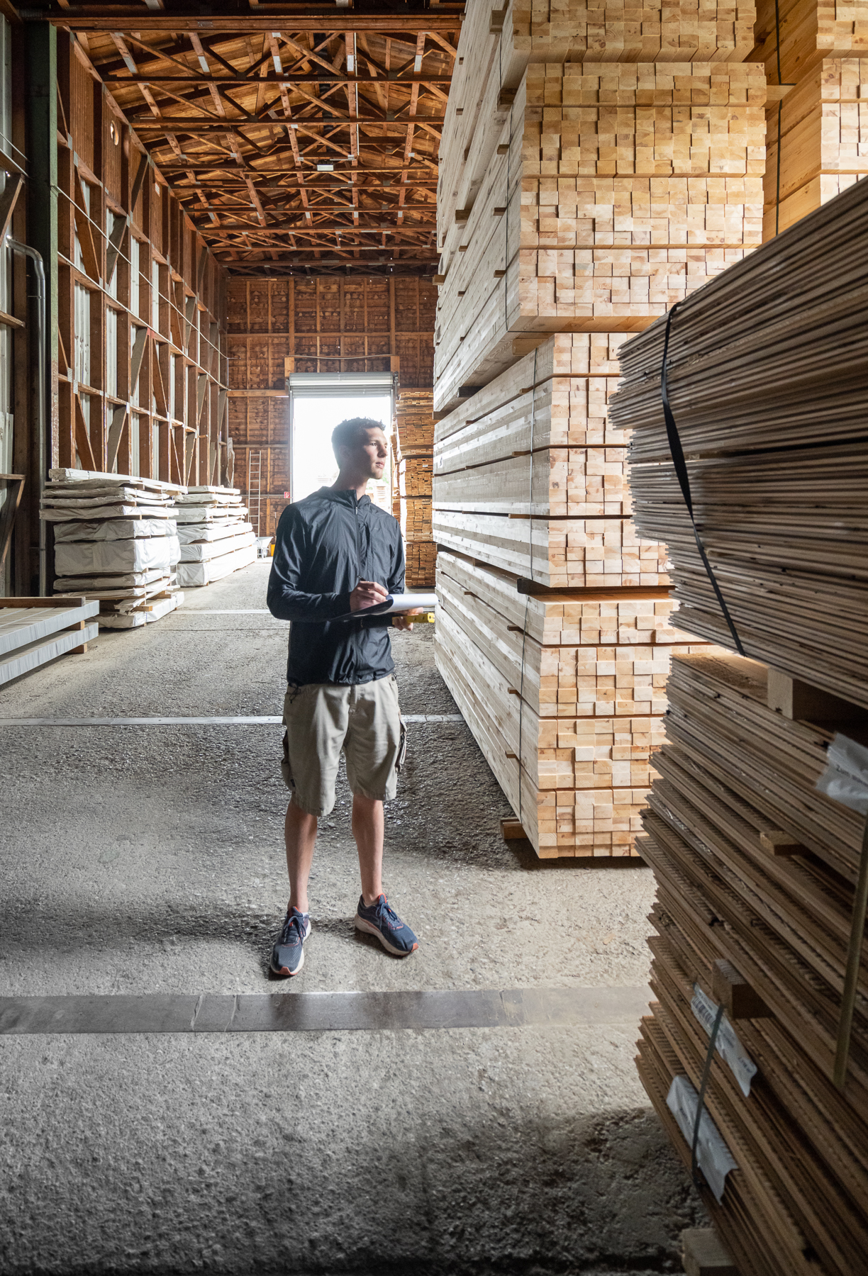 Ein Mitarbeiter inspiziert gestapelte, hell verarbeitete Holzbretter in einem großen Lagerhaus. Er hält Klemmbrett und Stift in der Hand und blickt prüfend auf die Holzstapel, die bis unter das Dach reichen.