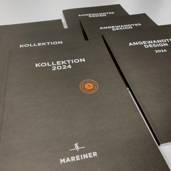 Mehrere dunkelbraune Broschüren mit der Aufschrift „KOLLEKTION 2024“ und „ANGEWANDTES DESIGN 2024“ von Mareiner Holz liegen auf einem Tisch. Das Titelblatt zeigt das Logo und ein Siegel in Orange.