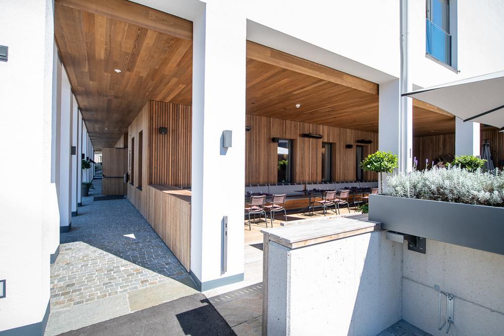 Blick auf den Durchgang und die angrenzende Terrasse des Hotel Restaurant zum Oxn. Die Fassade und Decke bestehen aus Mareiner Holz in Gardasee-Ausführung. Helle Putzflächen und Natursteinpflaster bilden einen eleganten Kontrast zum warmen Holz.