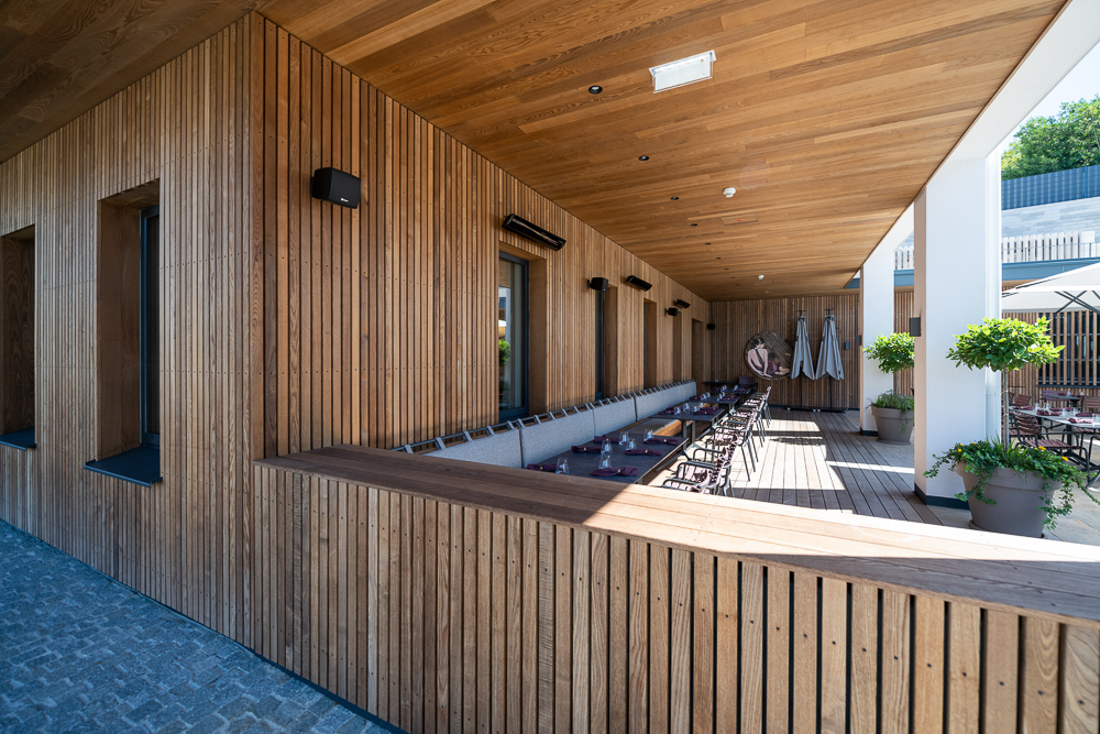 Außenansicht des überdachten Terrassenbereichs im Hotel Restaurant zum Oxn. Die vertikale Holzfassade und Decke in Mareiner Gardasee-Ausführung sorgen für eine warme, elegante Optik. Gedeckte Tische, Polsterbänke und Pflanzkübel laden zum Verweilen ein.