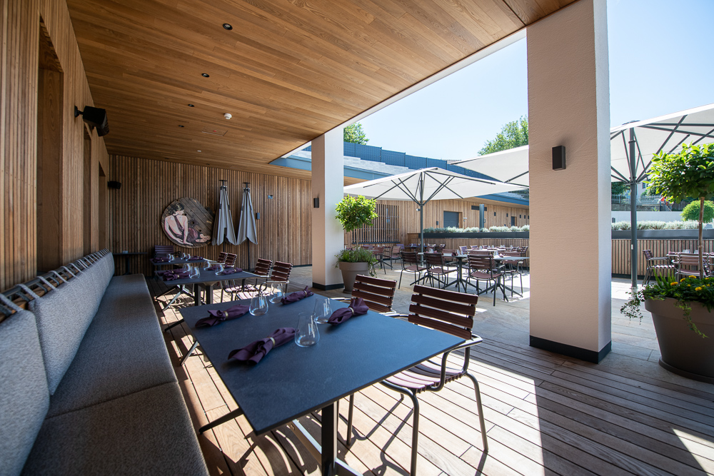 Teilweise überdachte Terrasse im Hotel Restaurant zum Oxn mit Holzfassade und Decke in Mareiner Gardasee-Ausführung. Sitzbänke mit Polsterung, gedeckte Tische, Sonnenschirme und üppige Pflanzkübel schaffen eine einladende Atmosphäre im Freien.
